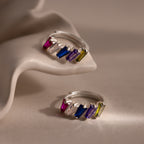 Two Multiple Tapered Birthstone Huggies in Sterling Silver, featuring rectangular multicolored gemstones, are displayed on a soft beige, sculptural surface.