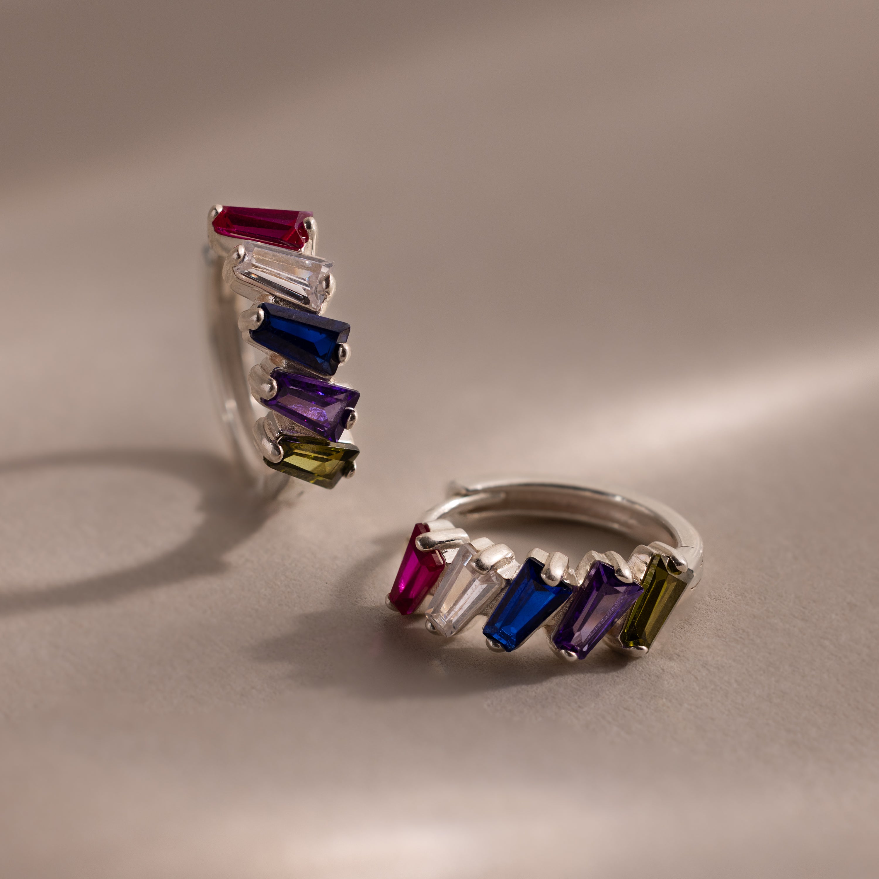 The Multiple Tapered Birthstone Huggies in Sterling Silver feature two silver hoops adorned with tapered gemstones in pink, white, blue, purple, and green—ideal for adding a playful art deco flair to any look.