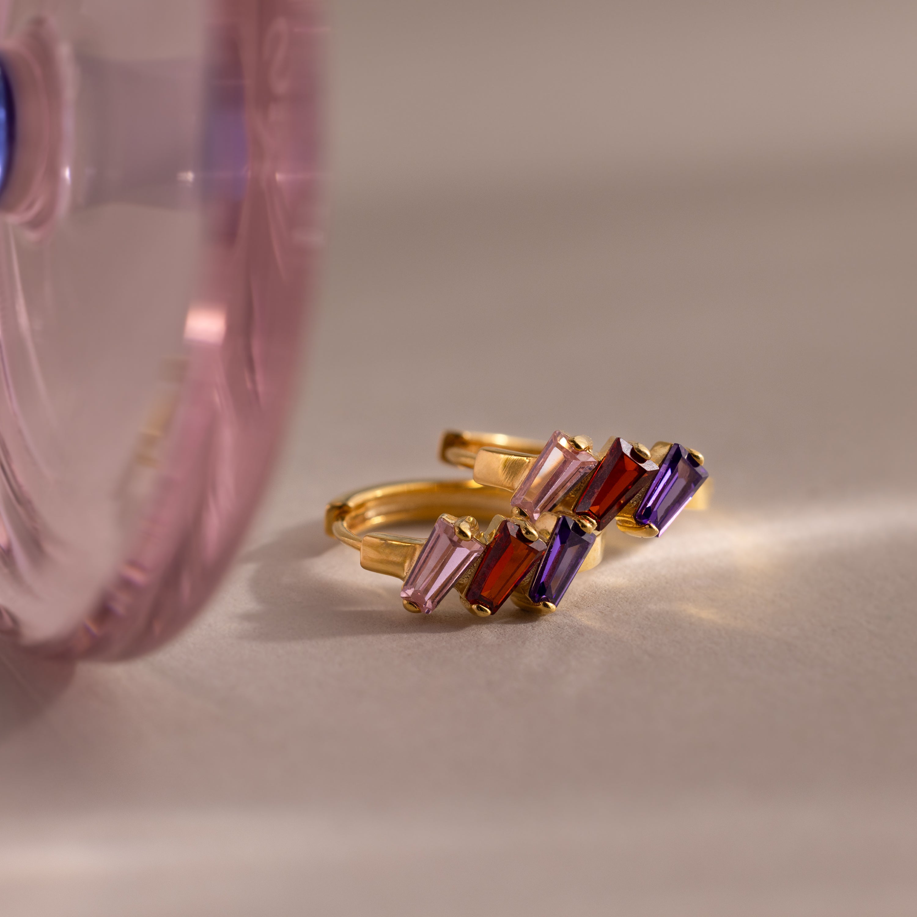 The Multiple Tapered Birthstone Huggies in 18K Gold feature pink, red, and purple gemstones, displayed beside a pink glass accent on a beige surface—an elegant, modern statement jewelry piece.