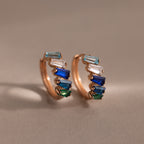 The Multiple Tapered Birthstone Huggies in Rose Gold feature rectangular blue, green, and clear gemstones in an Art Deco style, creating colorful statement earrings displayed on a neutral background.