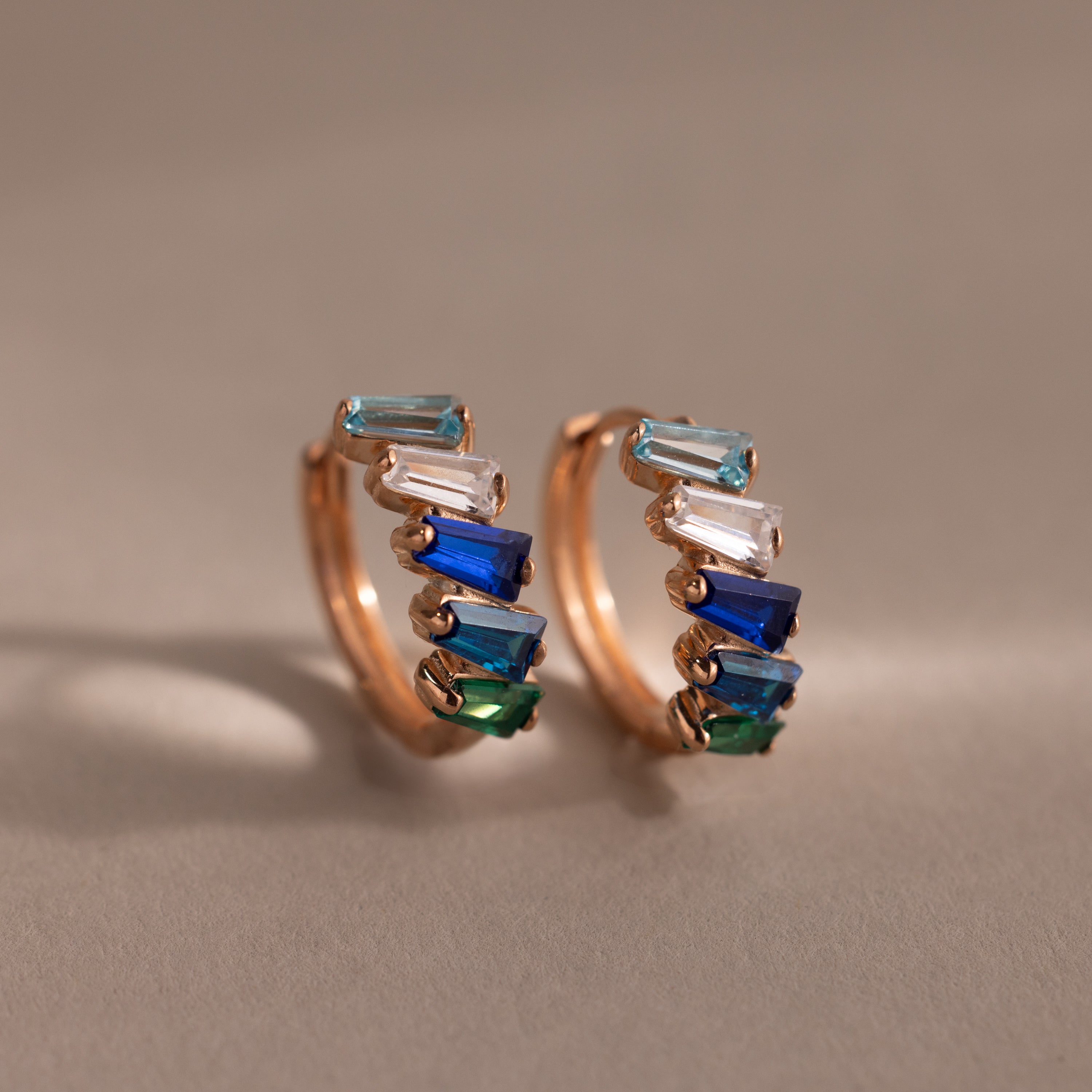 The Multiple Tapered Birthstone Huggies in Rose Gold feature rectangular blue, green, and clear gemstones in an Art Deco style, creating colorful statement earrings displayed on a neutral background.
