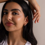 Woman with long dark hair wearing a white top and Multiple Tapered Birthstone Huggies in 18K Gold, smiling softly with hand raised above her head, embodying the allure of modern statement jewelry.