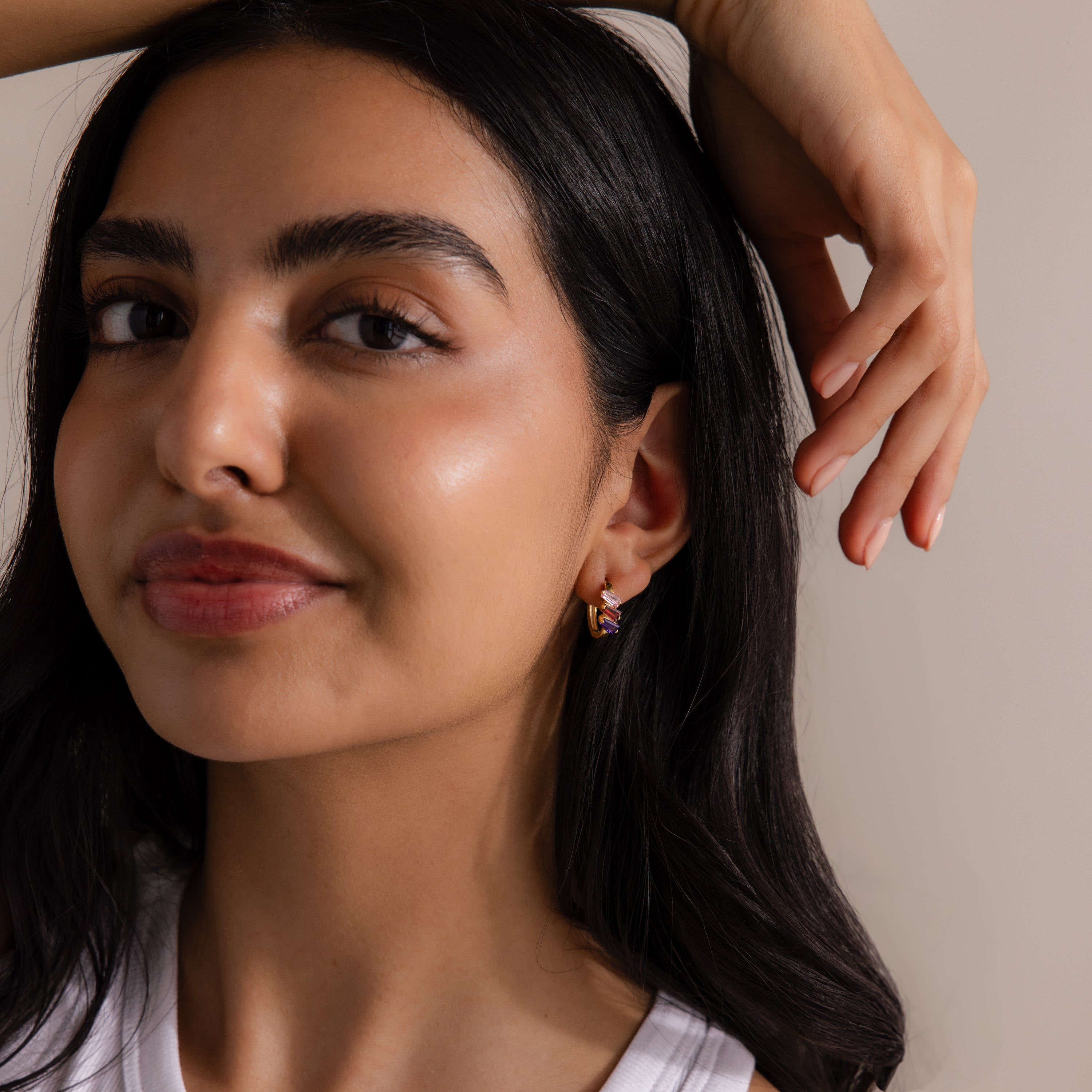 Woman with long dark hair wearing a white top and Multiple Tapered Birthstone Huggies in 18K Gold, smiling softly with hand raised above her head, embodying the allure of modern statement jewelry.