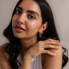 Woman with long dark hair wearing modern statement jewelry—Multiple Tapered Birthstone Huggies in gold—and a white top, resting her chin on her hand.