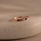 The Woven Multiple Birthstone Ring in Rose Gold features a vintage-inspired gold band set with five small red, purple, yellow, clear, and blue gemstones, displayed on a beige surface.