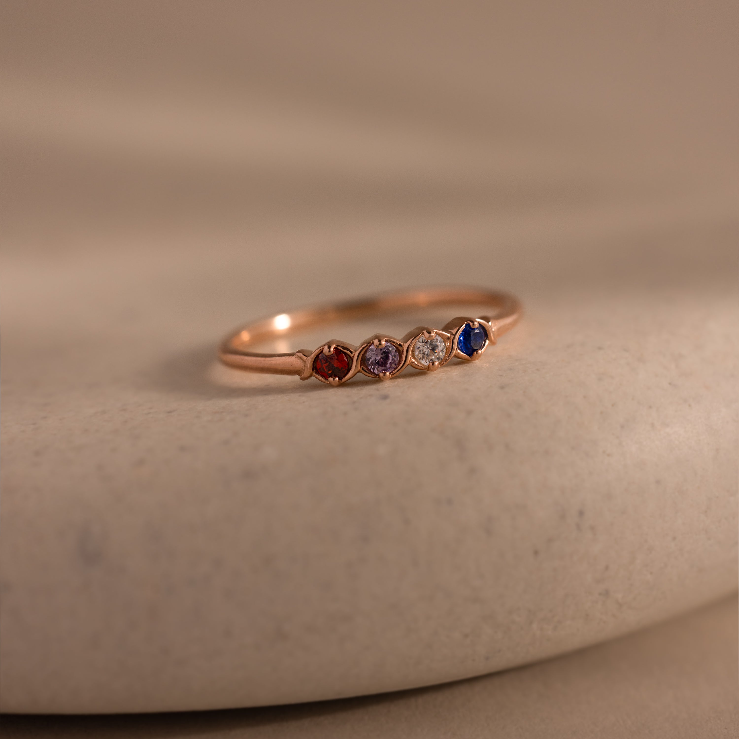 The Woven Multiple Birthstone Ring in Rose Gold features a vintage-inspired gold band set with five small red, purple, yellow, clear, and blue gemstones, displayed on a beige surface.