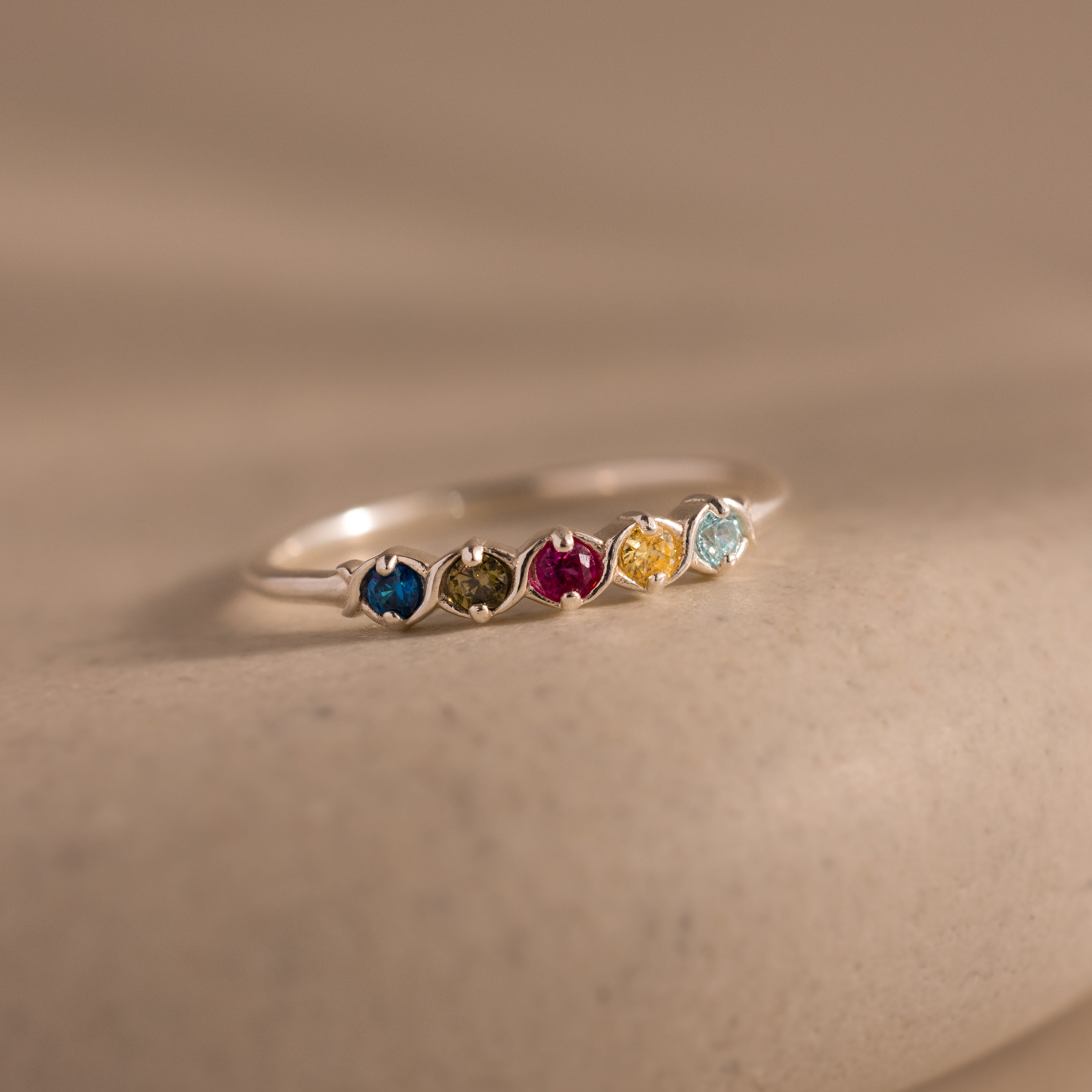 The Woven Multiple Birthstone Ring in Sterling Silver features five vibrant gemstones set in a delicate band, showcasing vintage-inspired elegance on a neutral surface.