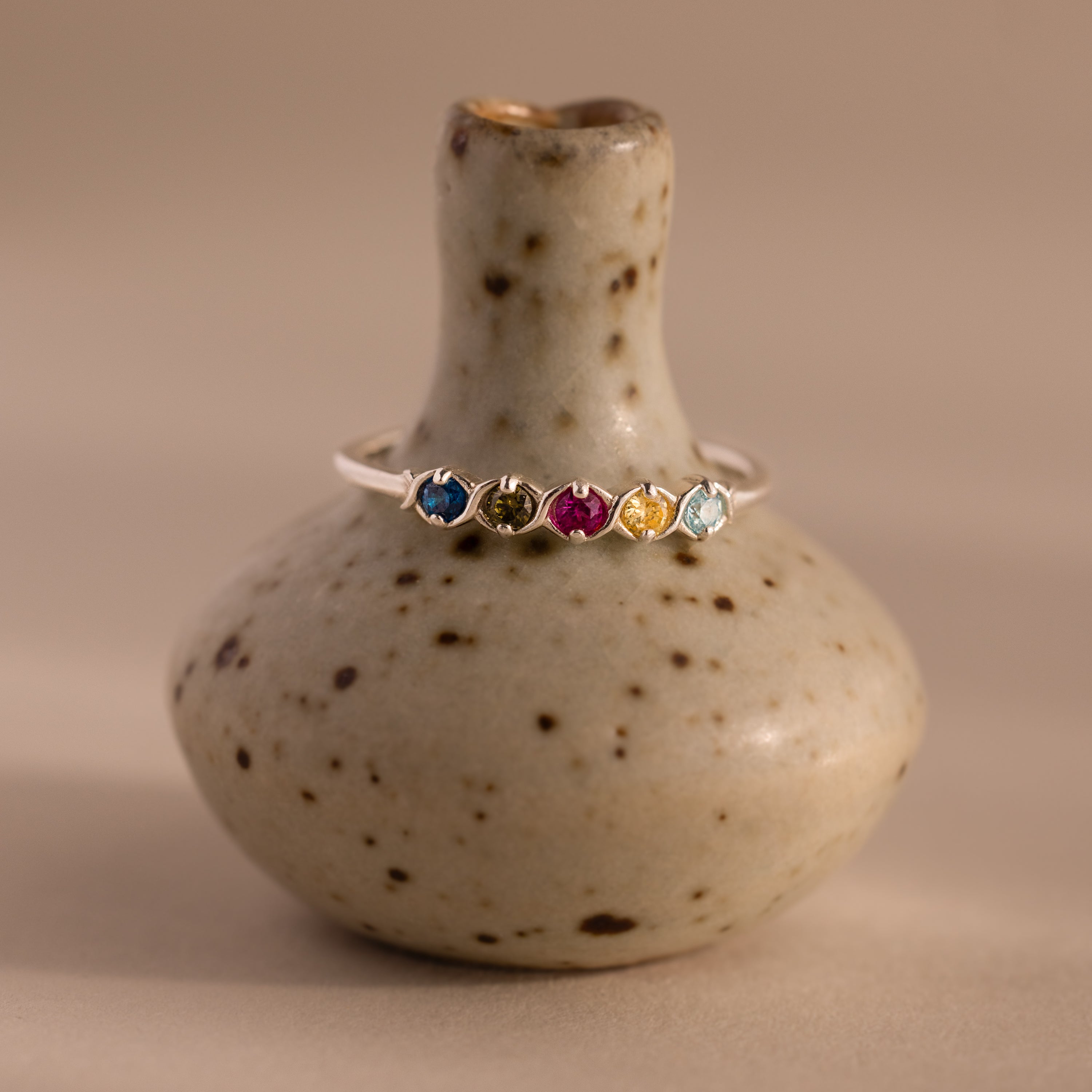 The Woven Multiple Birthstone Ring in Sterling Silver showcases five vibrant gemstones, arranged in a vintage-inspired design and displayed on a speckled ceramic vase.
