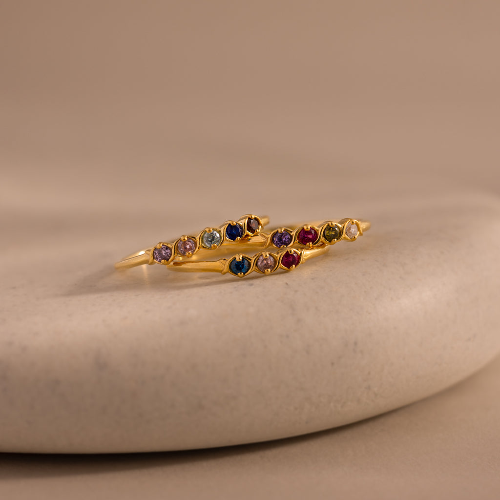 The Woven Multiple Birthstone Ring in 18K Gold features colorful gemstones, elegantly displayed on a smooth beige stone surface.