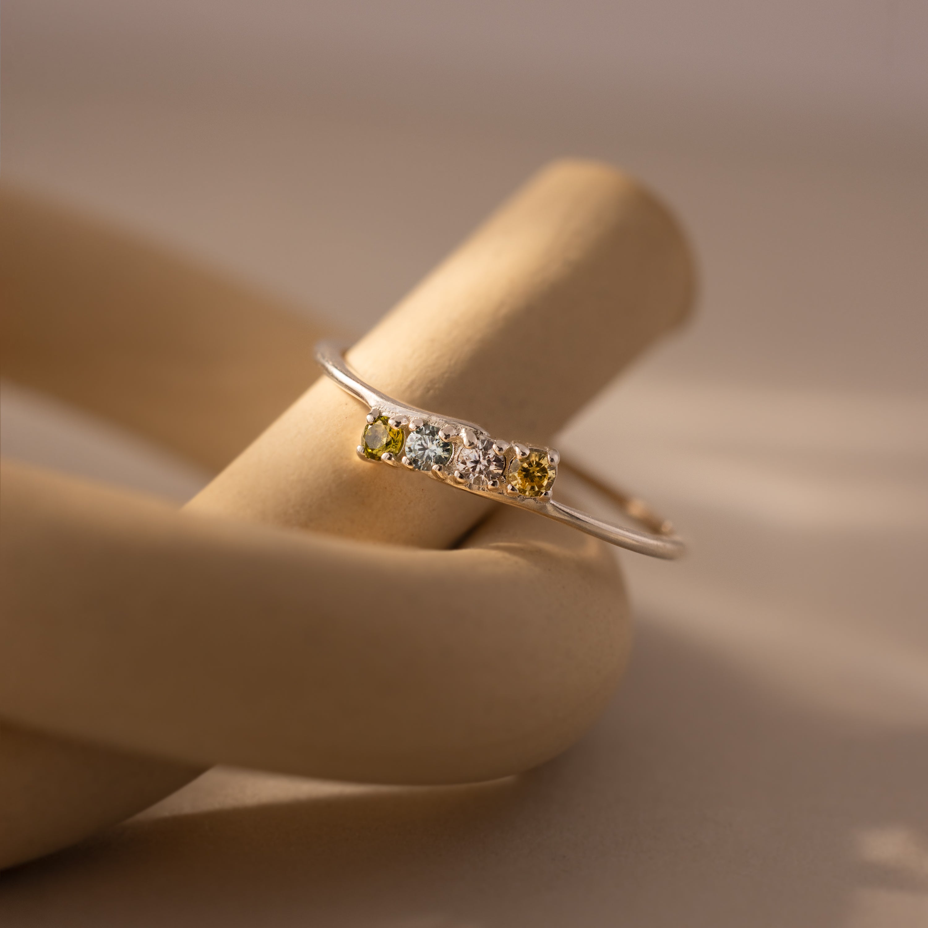 The Multiple Birthstone Curve Ring features a gold curved band with four yellow and clear gemstones, elegantly showcased on a beige sculptural surface.