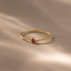 The Multiple Birthstone Curve Ring in 18K Gold, featuring a subtle ruby gemstone, is elegantly displayed on a soft beige surface.