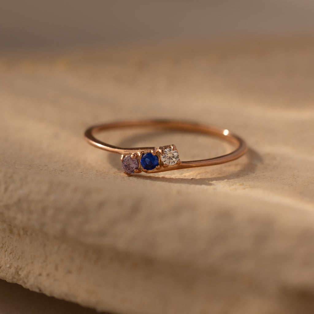 The Multiple Birthstone Curve Ring in Rose Gold features three petite stones—pink, blue, and clear—elegantly set on a beige textured surface.