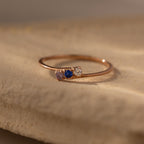The Multiple Birthstone Curve Ring in Rose Gold features three petite stones—pink, blue, and clear—elegantly set on a beige textured surface.