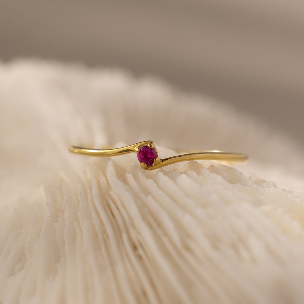 The Multiple Birthstone Curve Ring in 18K Gold featuring a small pink gemstone is displayed on a textured, creamy white surface.