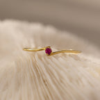 The Multiple Birthstone Curve Ring in 18K Gold featuring a small pink gemstone is displayed on a textured, creamy white surface.