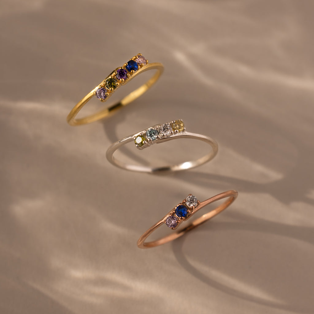 The Multiple Birthstone Curve Ring in Rose Gold features a delicate band with multicolored gemstones—perfect for stacking or wearing solo and beautifully showcased on a neutral surface.