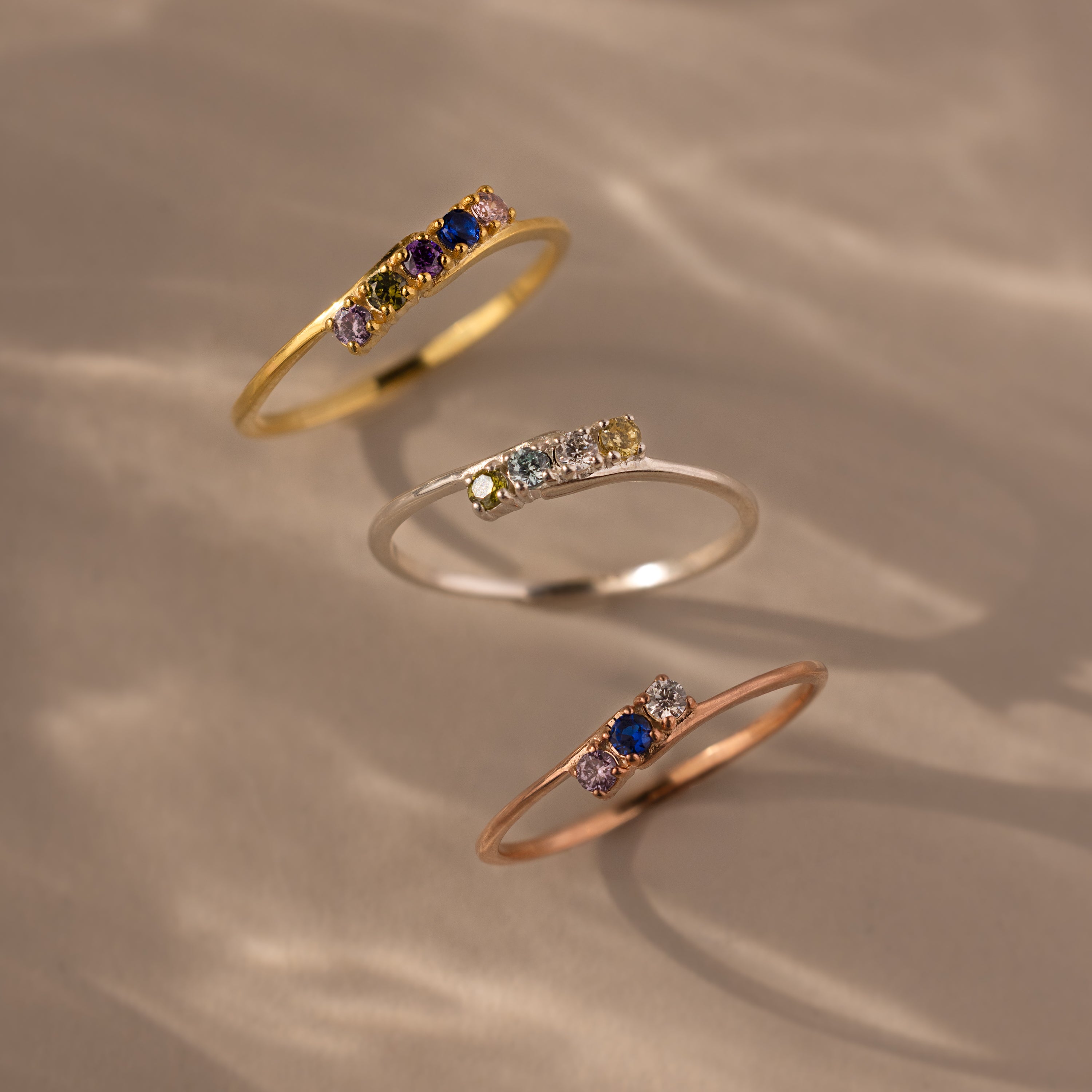 The Multiple Birthstone Curve Ring in Rose Gold features a delicate band with multicolored gemstones—perfect for stacking or wearing solo and beautifully showcased on a neutral surface.