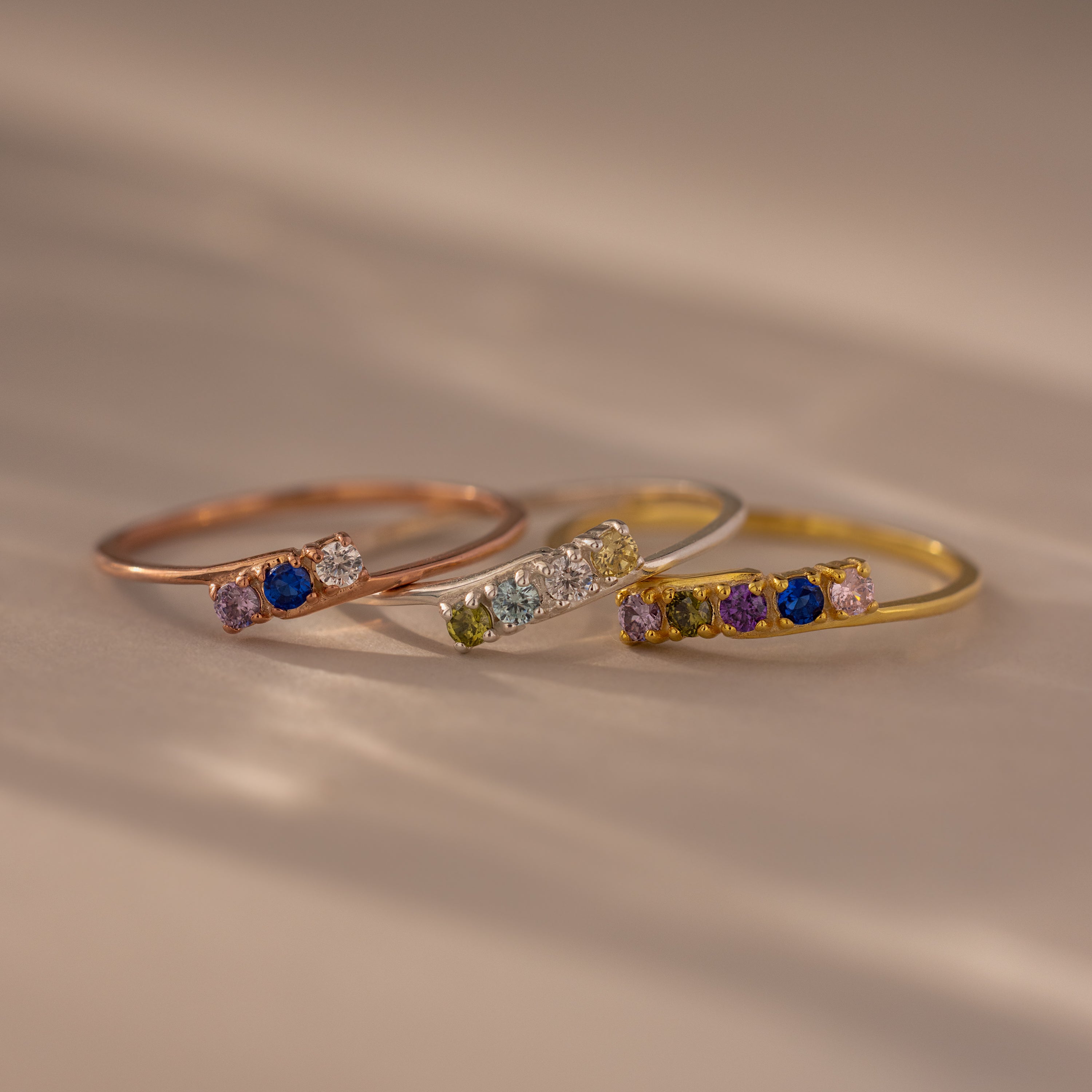 The Multiple Birthstone Curve Ring, available in rose gold, silver, and gold, features small colorful gemstones and rests elegantly on a soft surface.