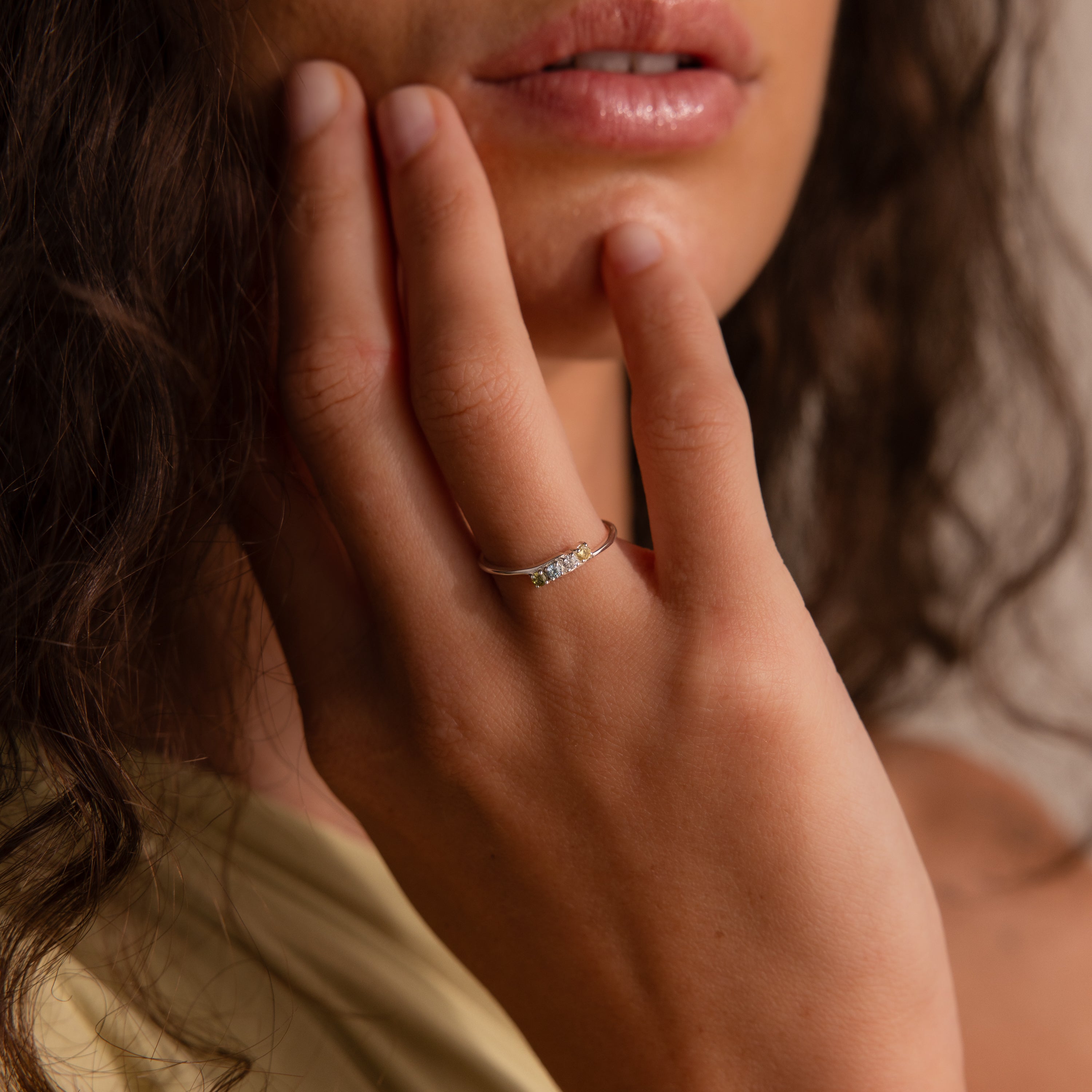 A woman touches her face, showcasing the Multiple Birthstone Curve Ring on her finger.