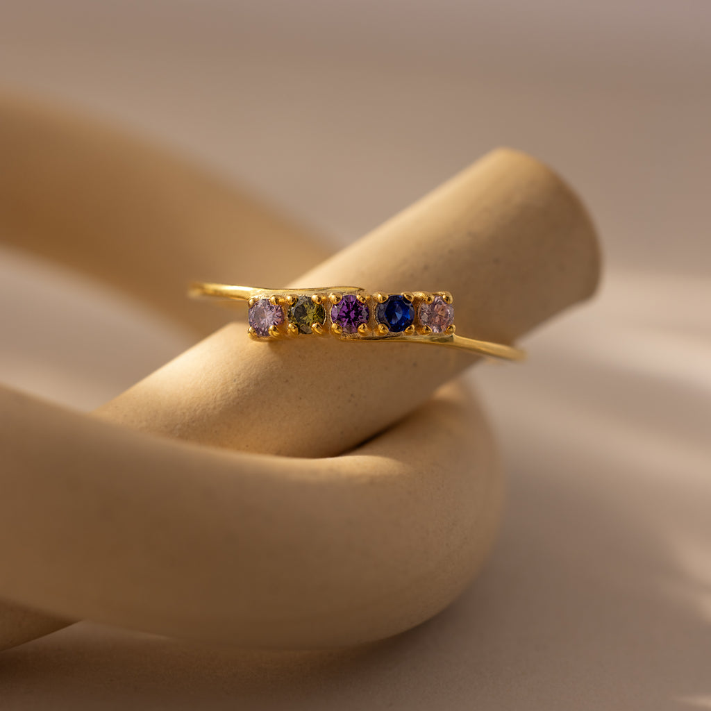 The Multiple Birthstone Curve Ring in 18K Gold, featuring vibrant gemstones, is displayed on a curved, beige ceramic stand.