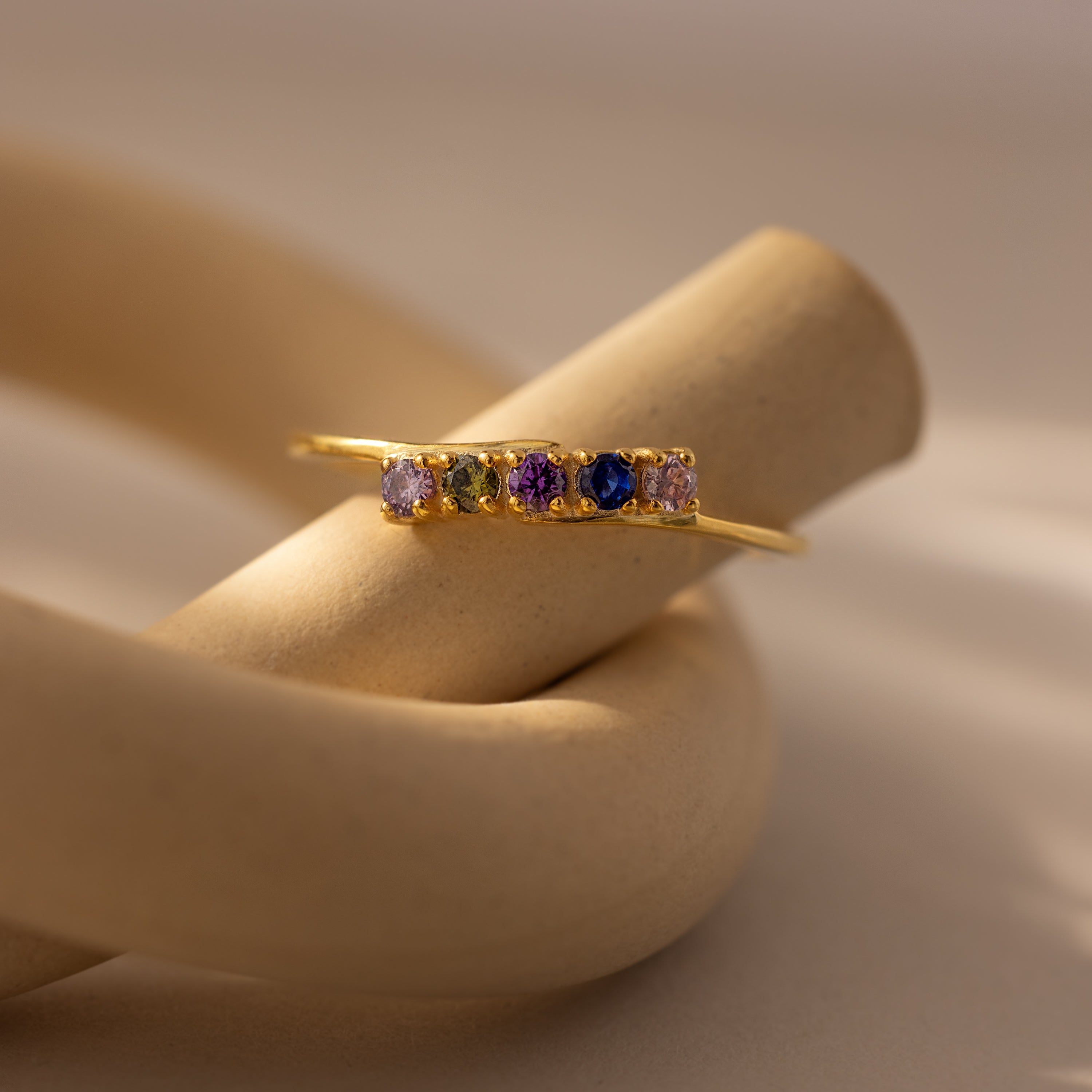 The Multiple Birthstone Curve Ring in 18K Gold, featuring vibrant gemstones, is displayed on a curved, beige ceramic stand.