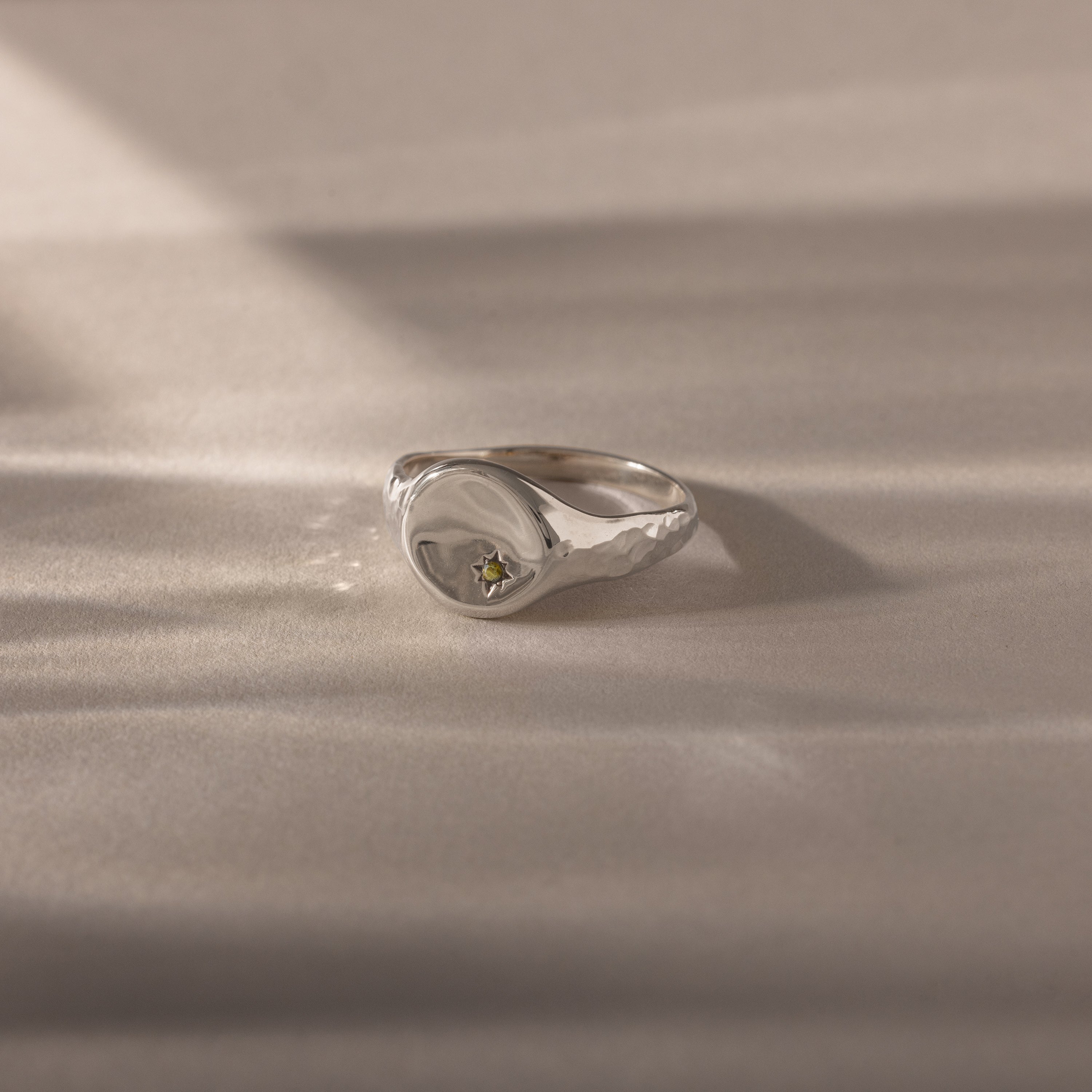 The North Star Peridot Signet Ring, a silver band featuring a small yellow peridot gem, is displayed as symbolic jewelry on a softly lit beige surface.