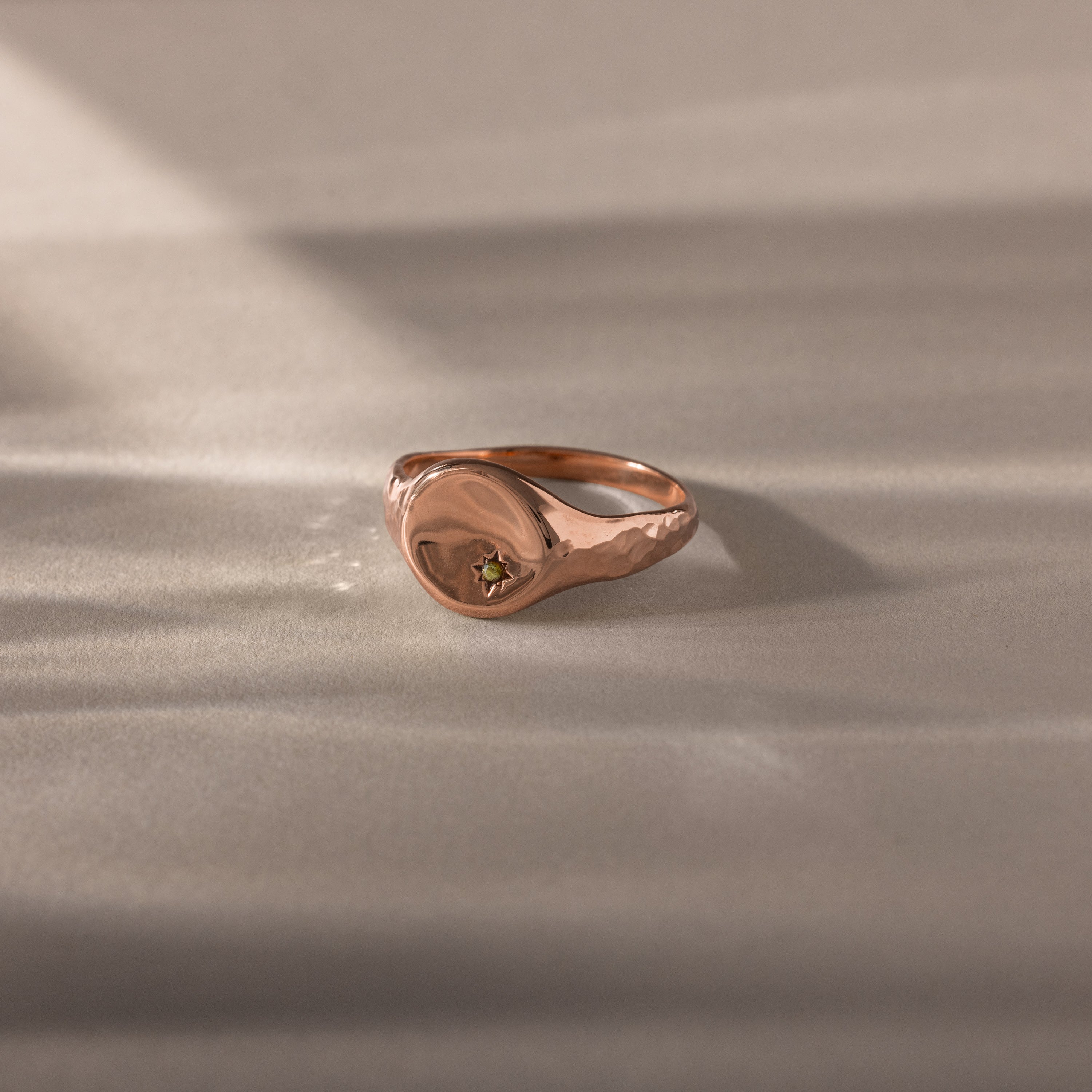 A symbolic jewelry piece, the North Star Peridot Signet Ring in rose gold features a small green peridot gem, elegantly displayed on a neutral, softly lit surface.