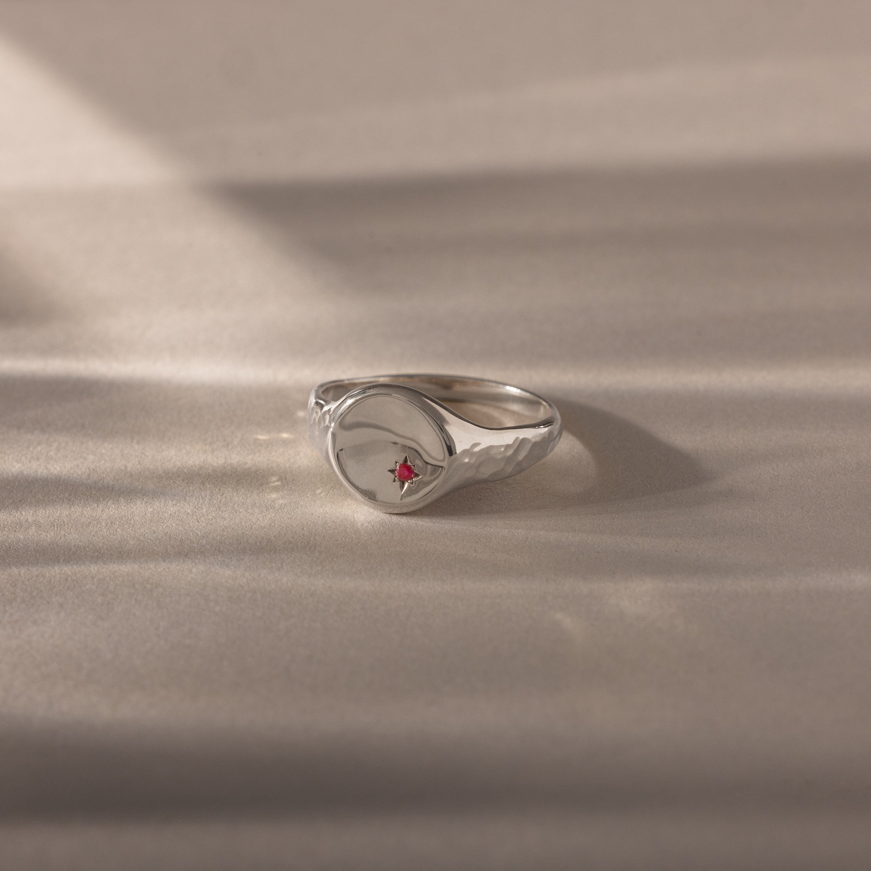The North Star Ruby Signet Ring is a symbolic silver jewelry piece featuring a central red gemstone, showcased on a soft beige surface.