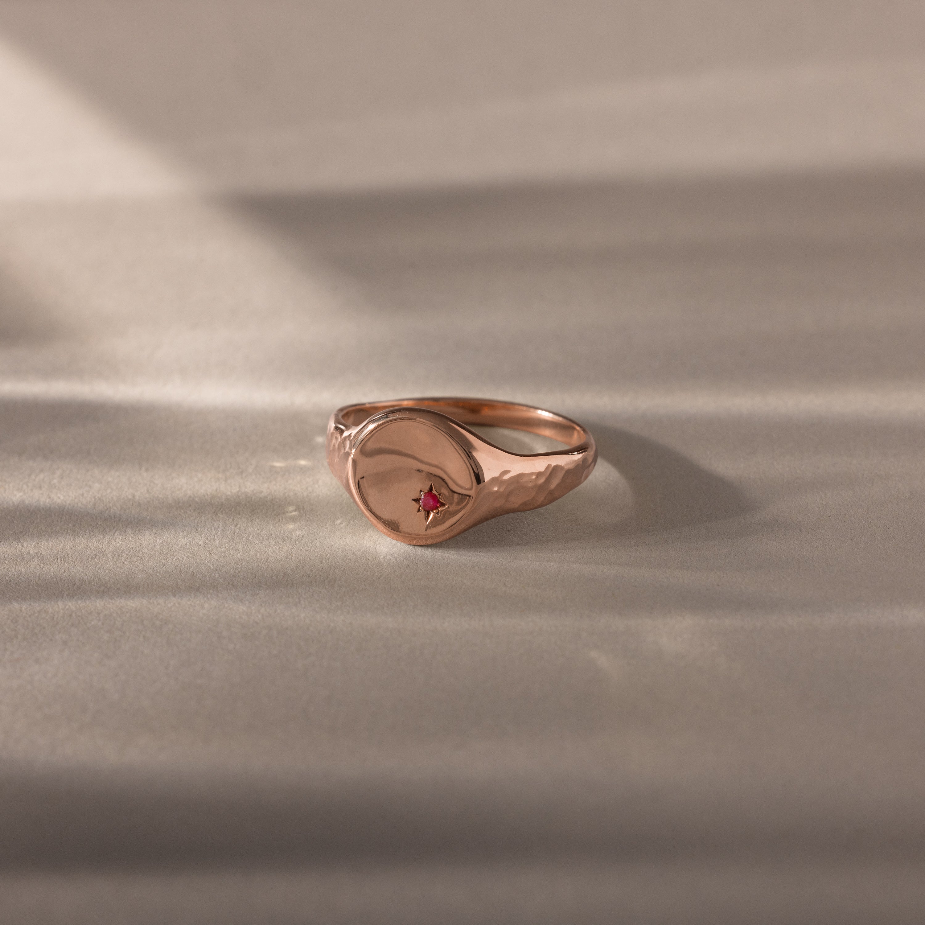 The North Star Ruby Signet Ring features a small red gemstone set in rose gold and is displayed on a neutral surface—a stunning piece of symbolic jewelry.