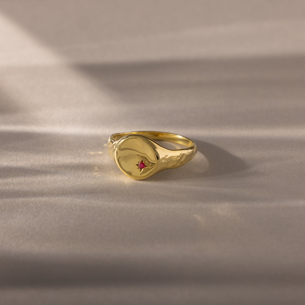 The North Star Birthstone Signet Ring in Ruby (July), a symbolic jewelry piece, is displayed on a softly lit beige surface.