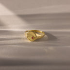 The North Star Birthstone Signet Ring in Ruby (July), a symbolic jewelry piece, is displayed on a softly lit beige surface.