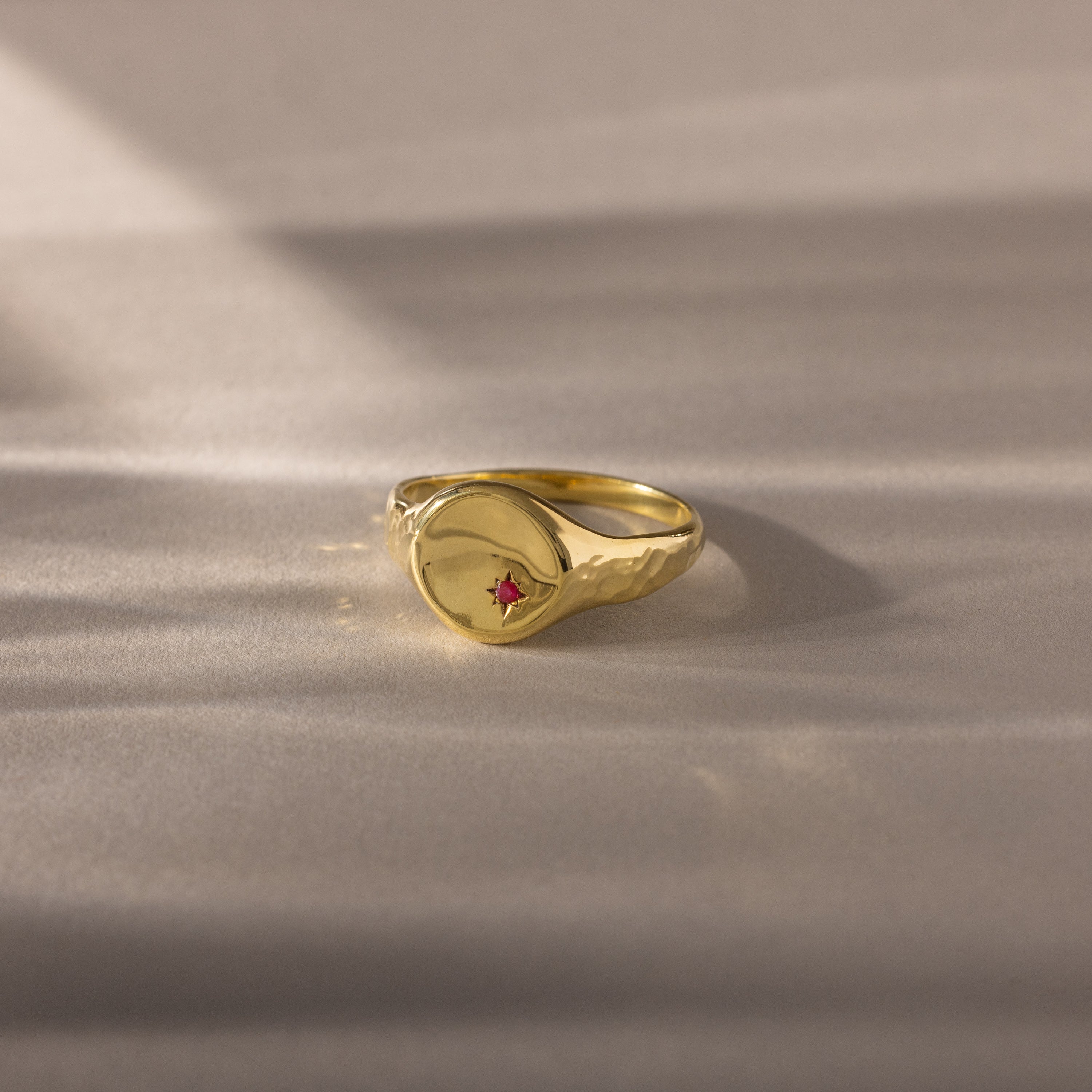 The North Star Birthstone Signet Ring in Ruby (July), a symbolic jewelry piece, is displayed on a softly lit beige surface.