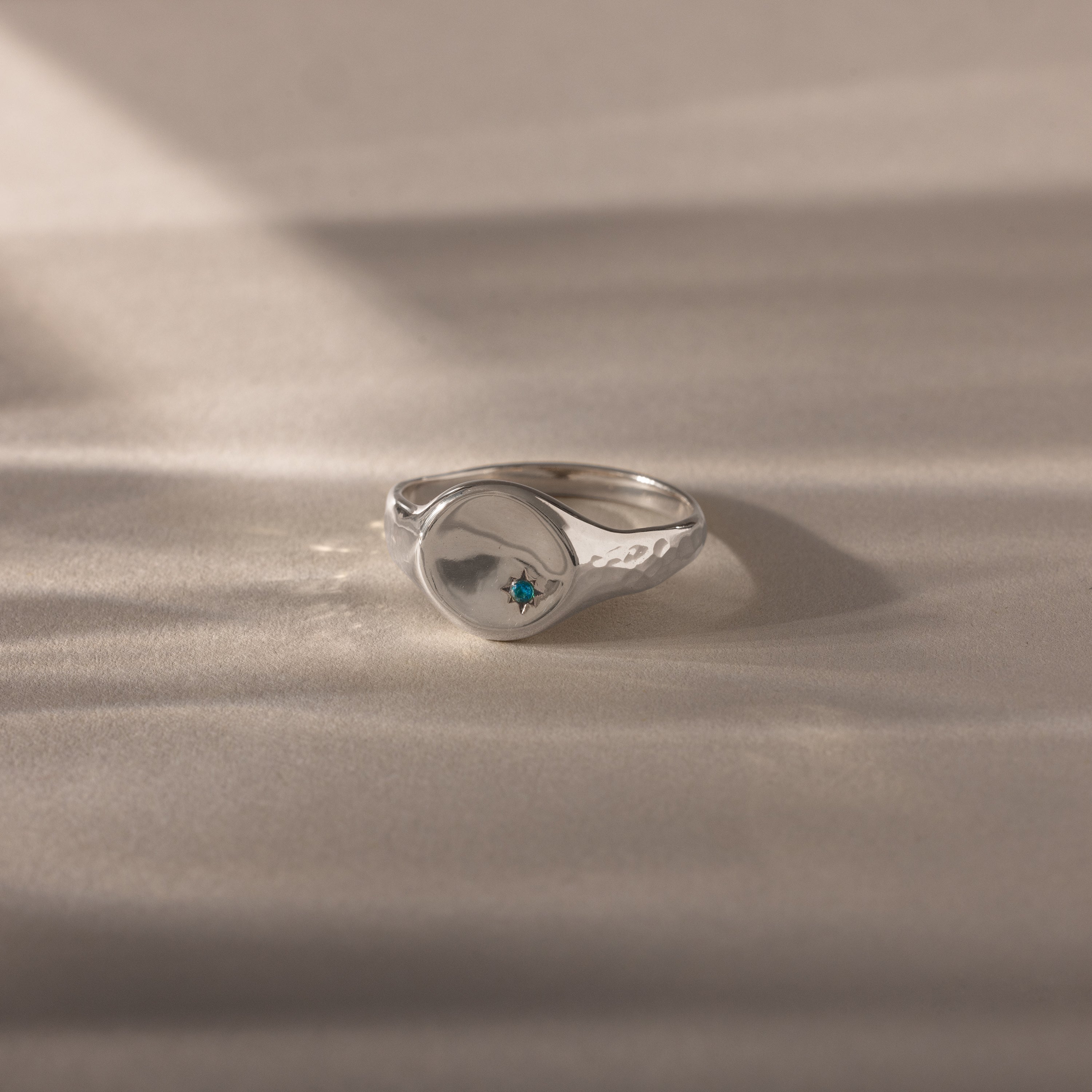 The North Star Zircon Signet Ring features a silver band with a small blue zircon gemstone at its center, displayed on a soft beige surface—an elegant and symbolic jewelry piece.