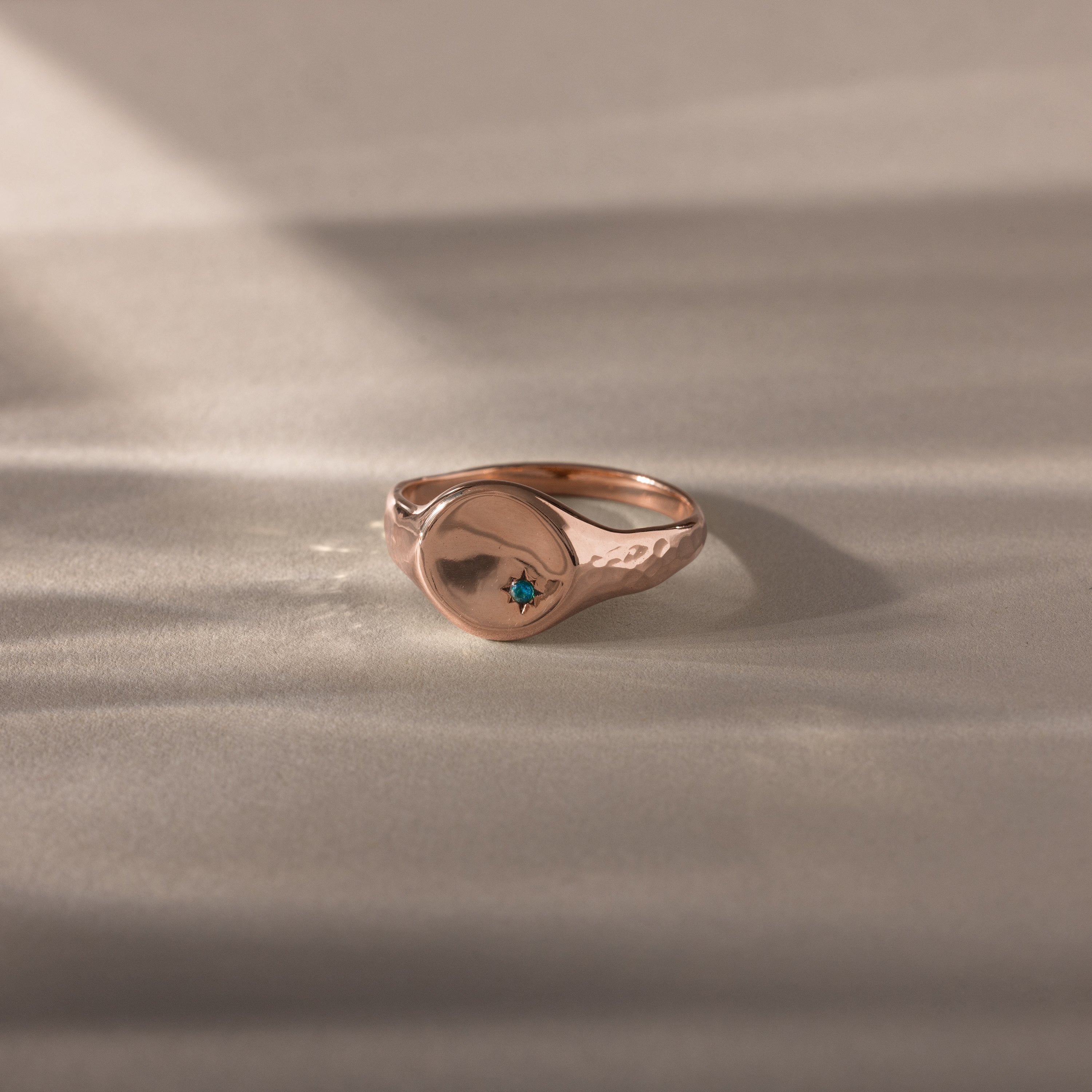 The North Star Zircon Signet Ring features a rose gold hammered finish and a central blue zircon gemstone, displayed on a light surface.