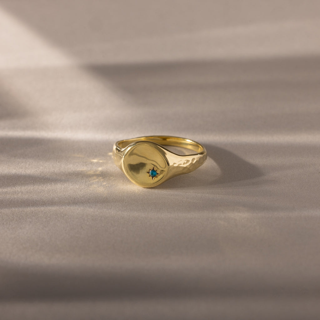 The North Star Birthstone Signet Ring, featuring a hammered gold finish and a central blue zircon for December birthdays, rests on a soft beige surface.