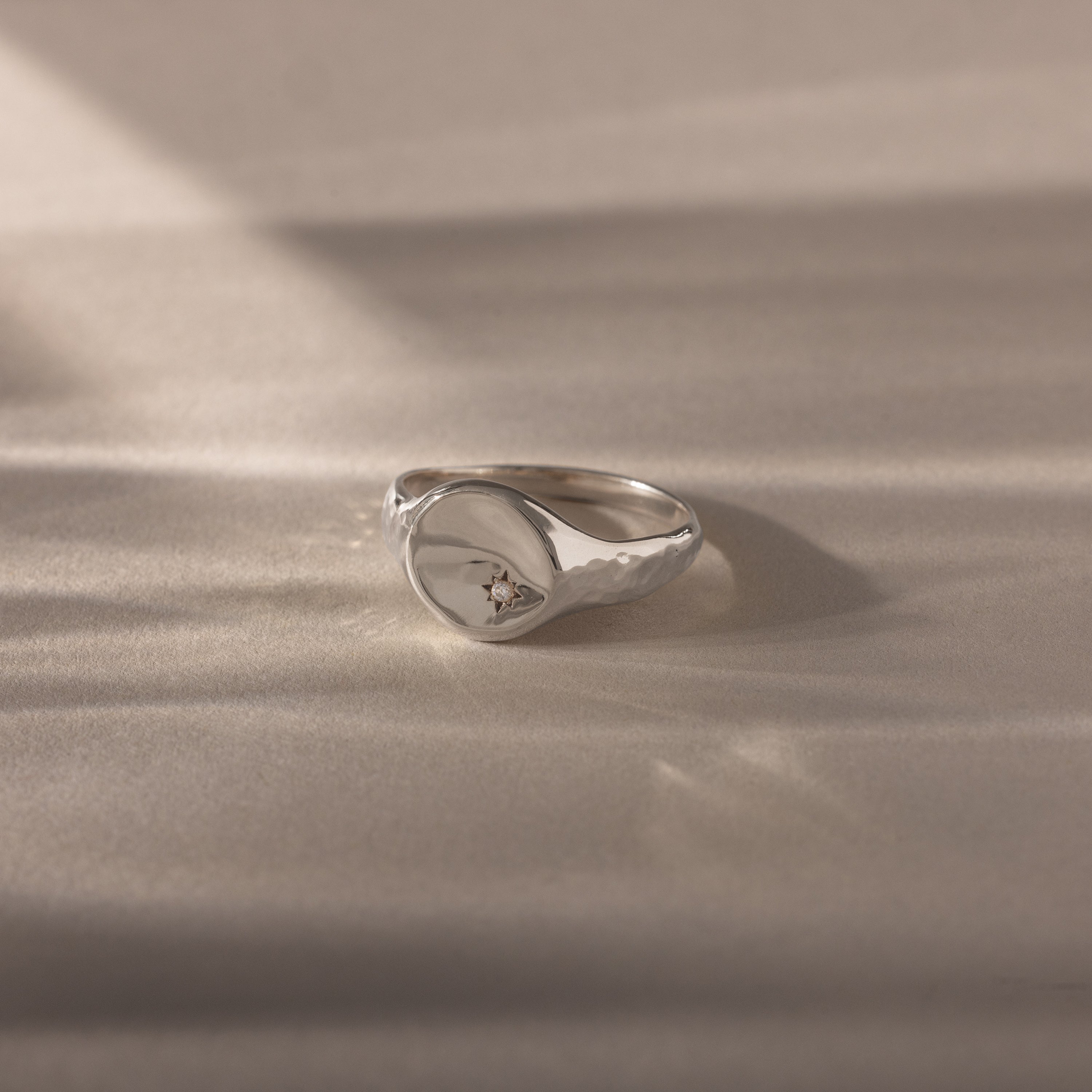 The North Star Diamond Signet Ring, crafted in silver, features a round, subtly hammered top with an off-center diamond accent, elegantly displayed on a soft beige surface.