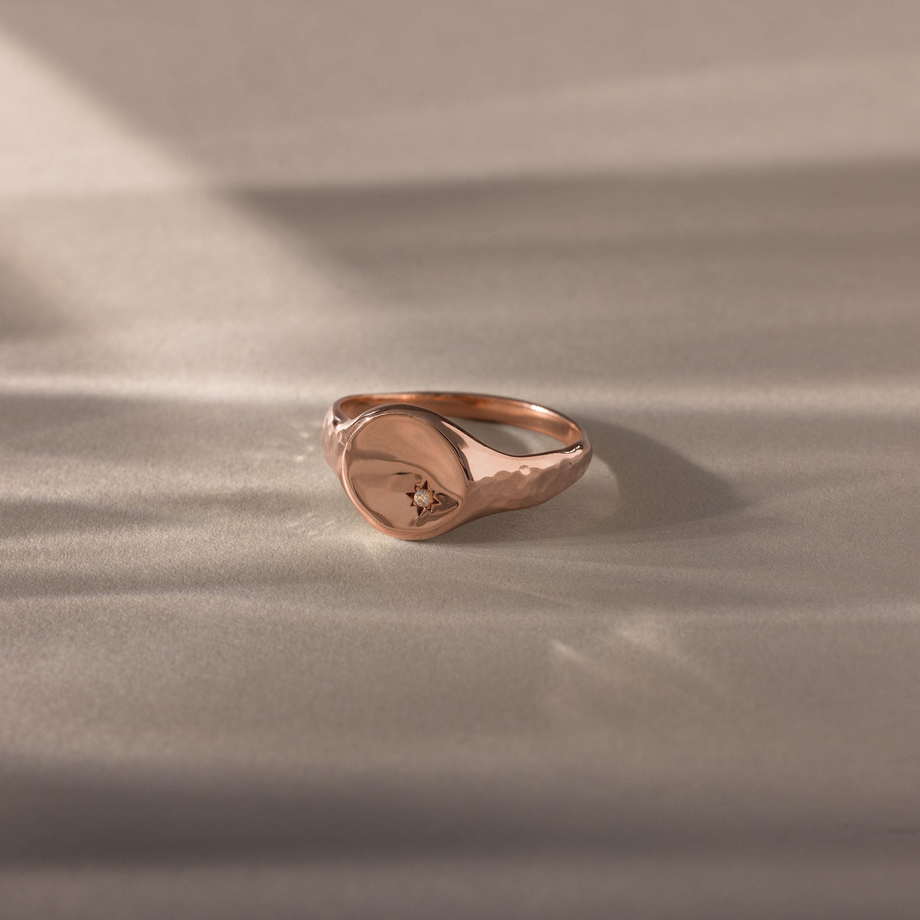 A symbolic jewelry piece, the North Star Diamond Signet Ring in rose gold with a small diamond detail rests on a soft, light-colored surface.