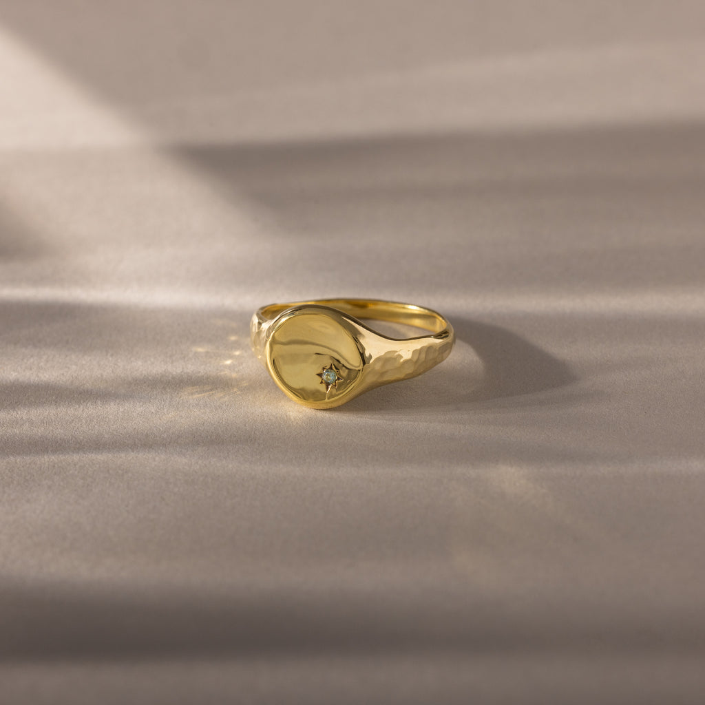 The North Star Birthstone Signet Ring in aquamarine (March) features a round face with a central gemstone, shown on a soft beige surface—an elegant, symbolic jewelry piece.