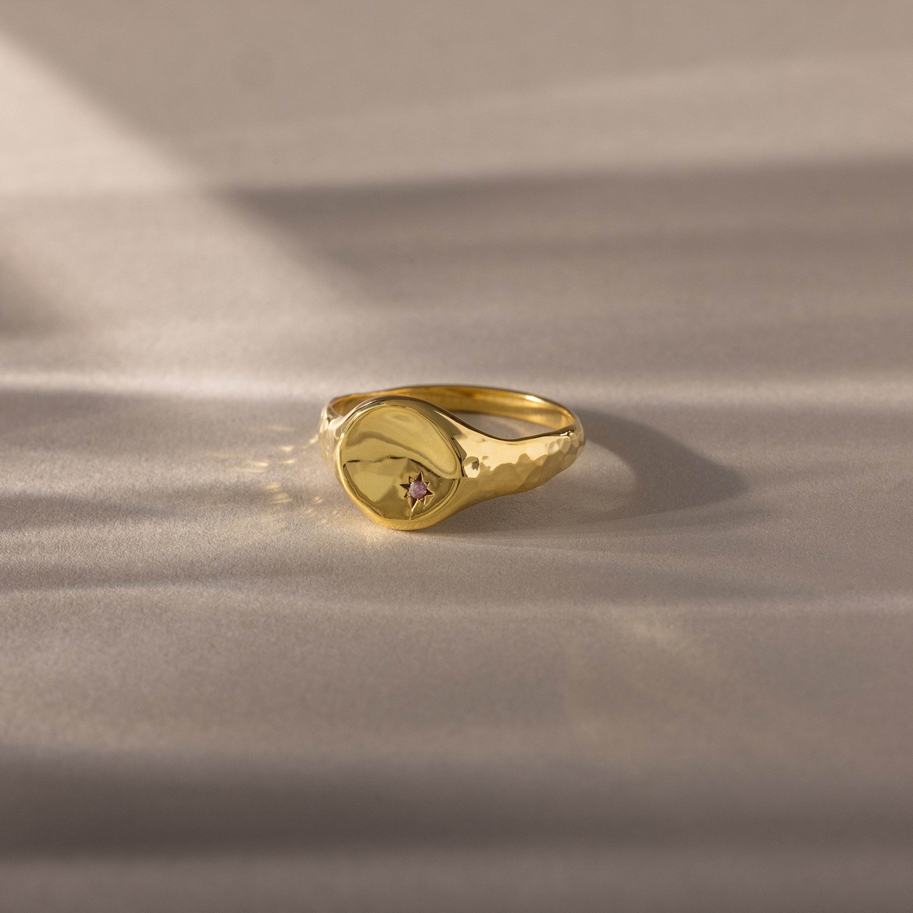 The North Star Alexandrite Signet Ring features a hammered gold band with a small pink alexandrite gemstone at its center, displayed on a soft surface.