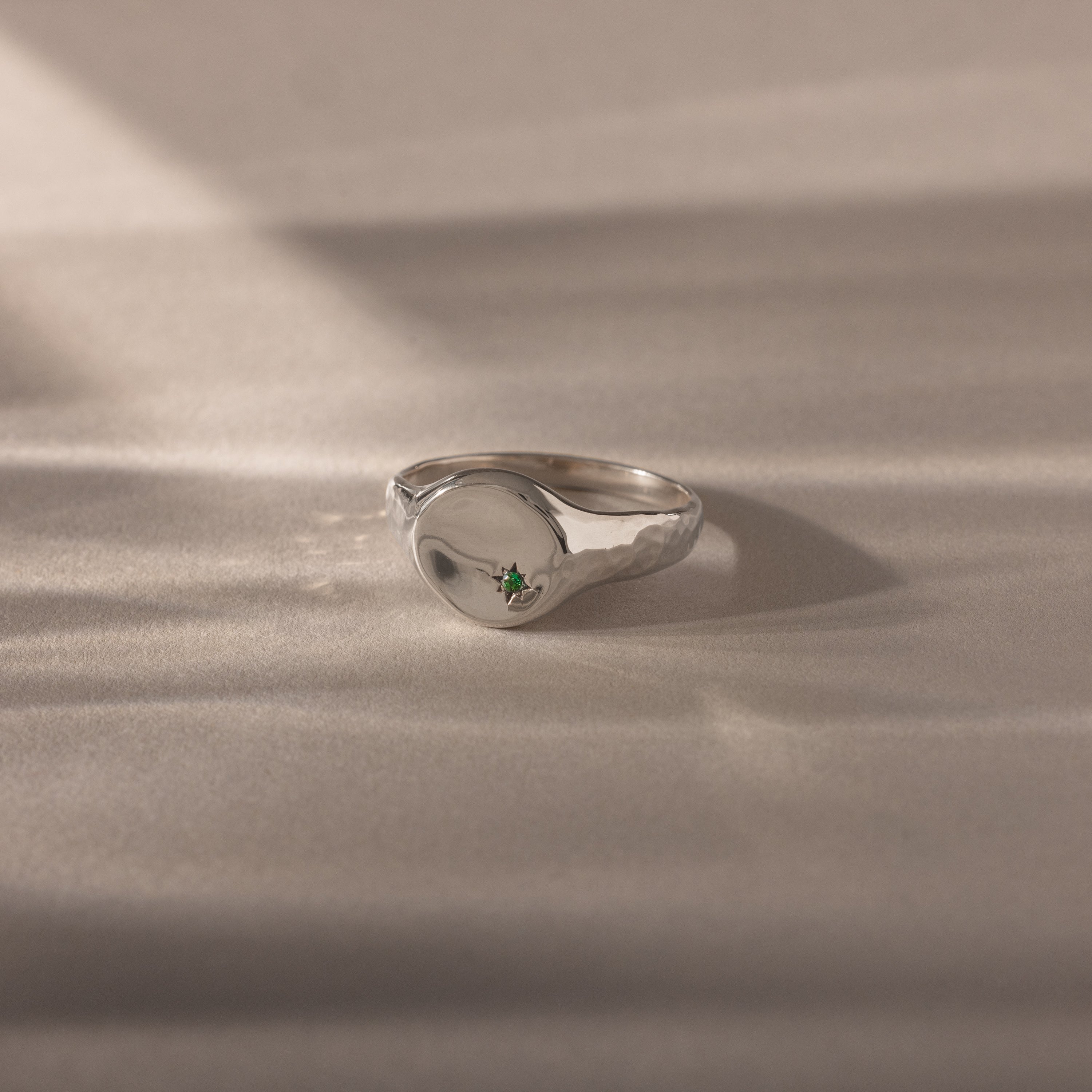 The North Star Emerald Signet Ring, featuring a silver band and a small green gemstone, is shown on a soft neutral surface with gentle shadows—an elegant piece of symbolic jewelry.