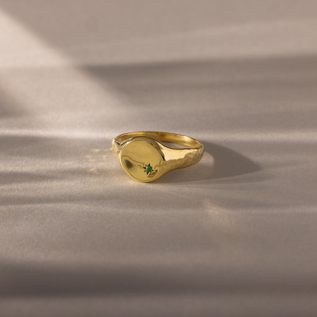 The North Star Birthstone Signet Ring in emerald (May) features a small green gemstone at its center and is showcased on a soft beige surface.