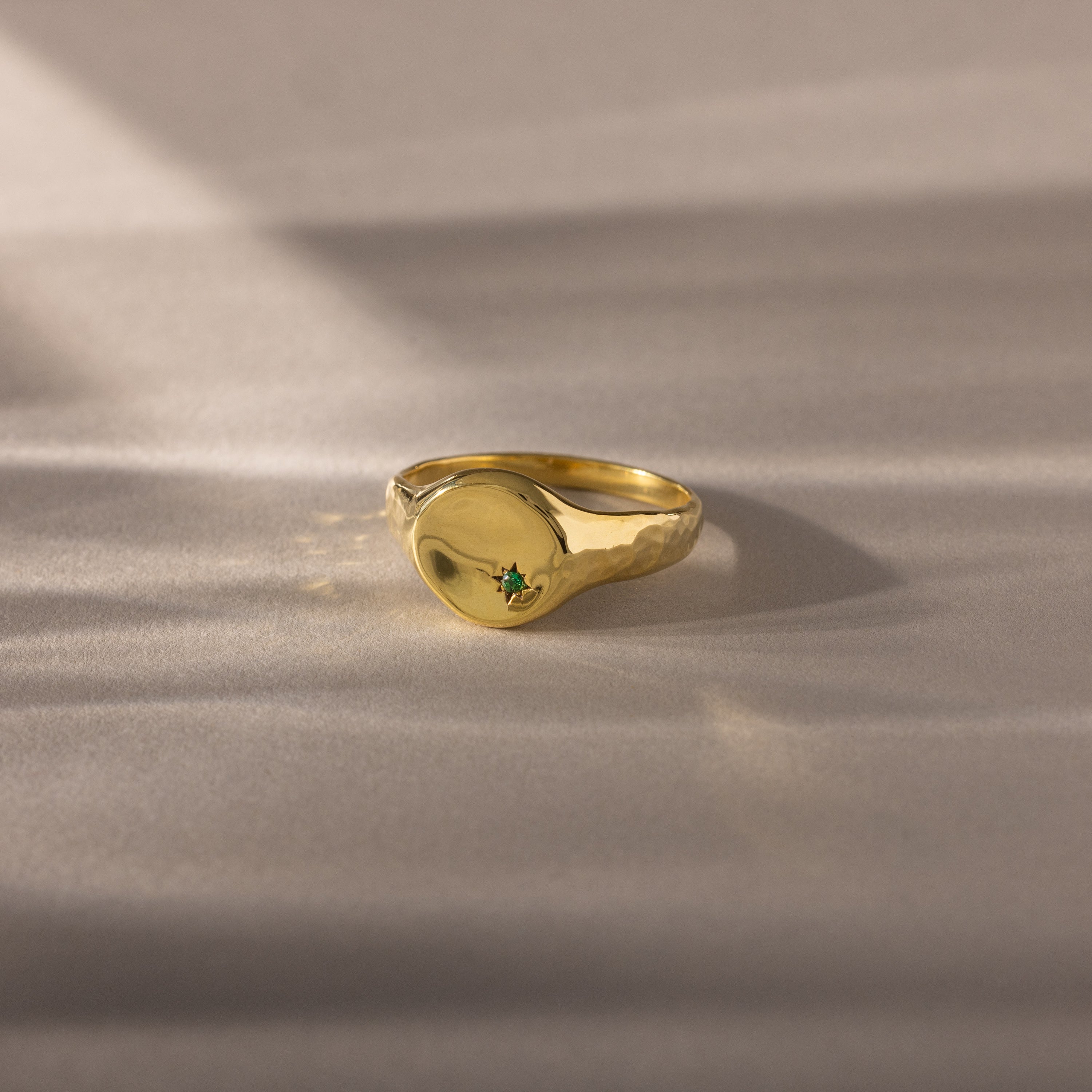 The North Star Birthstone Signet Ring in emerald (May) features a small green gemstone at its center and is showcased on a soft beige surface.