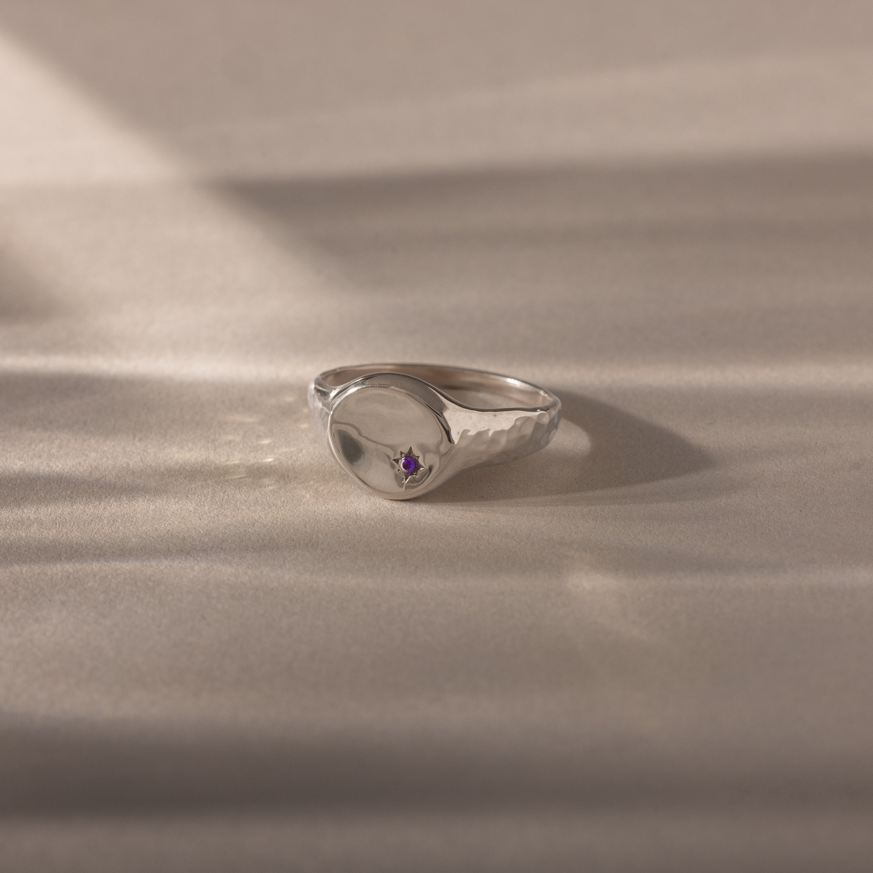 The North Star Amethyst Signet Ring, featuring a silver band and a small purple gemstone, is showcased on a soft, neutral background—a meaningful, symbolic jewelry piece.