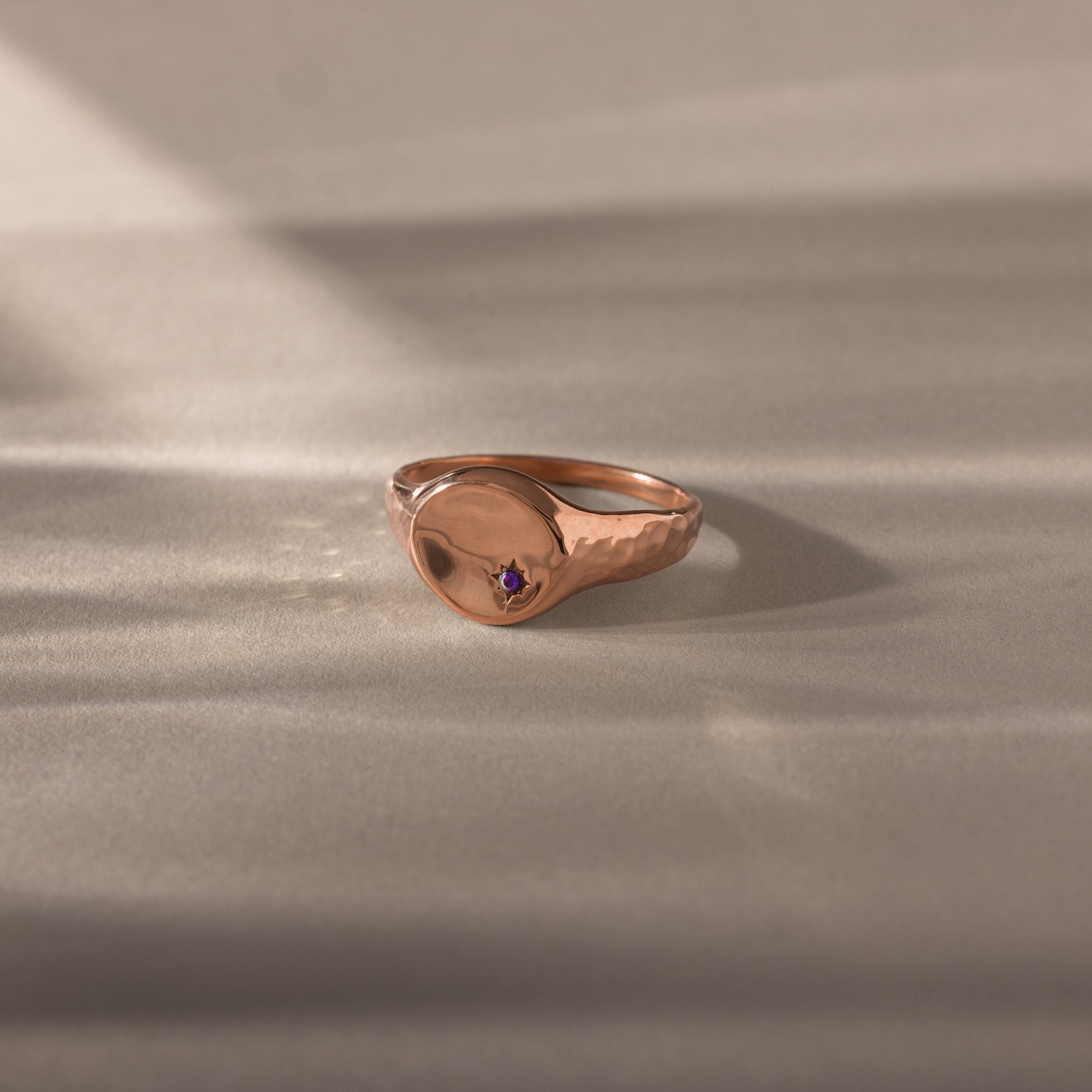 The North Star Amethyst Signet Ring in rose gold features a small purple gemstone, showcased on a light textured surface with soft shadows—an elegant piece of symbolic jewelry.