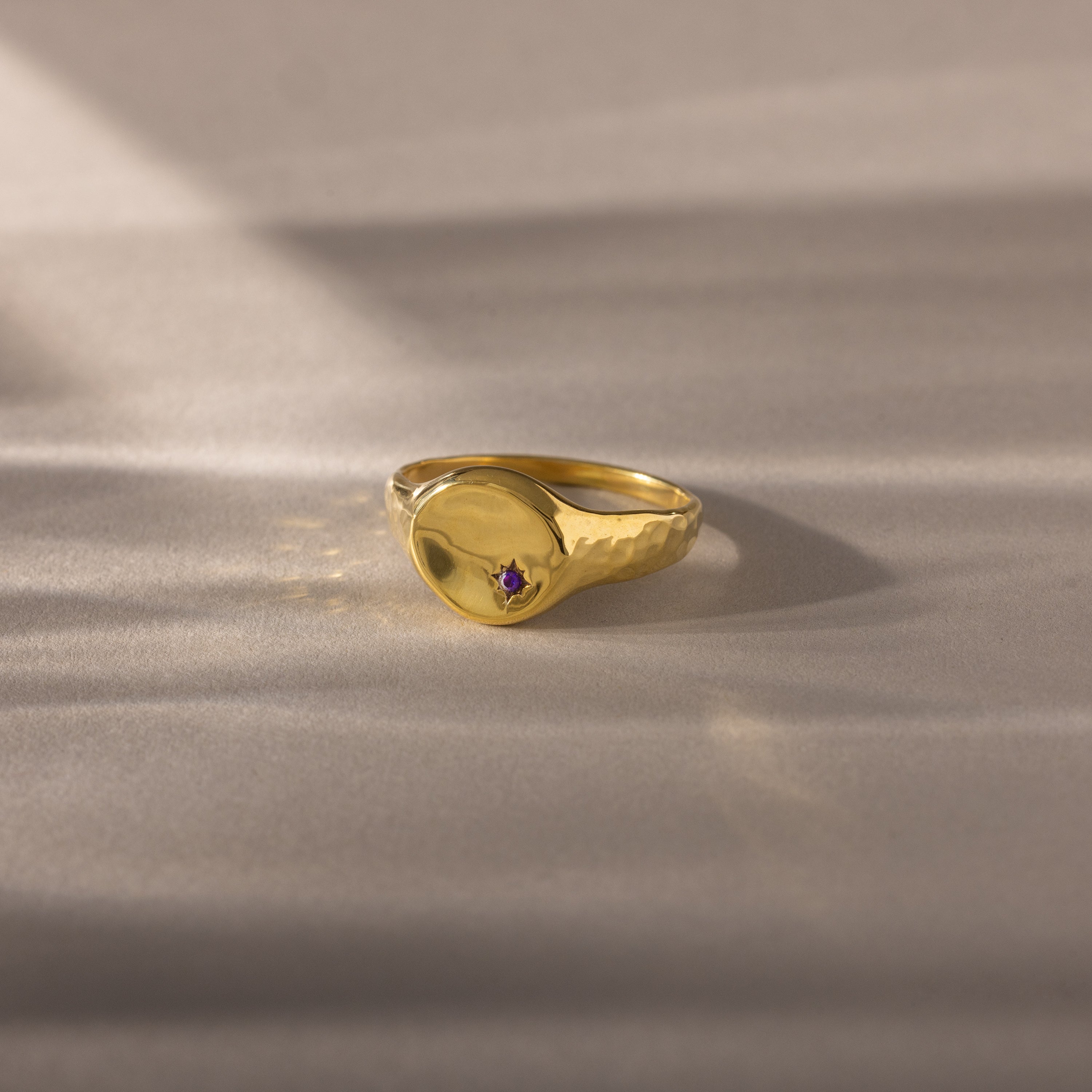 The North Star Amethyst Signet Ring features a hammered gold band with a small purple amethyst centerpiece, set on a soft beige surface—an ideal symbolic piece for everyday wear.