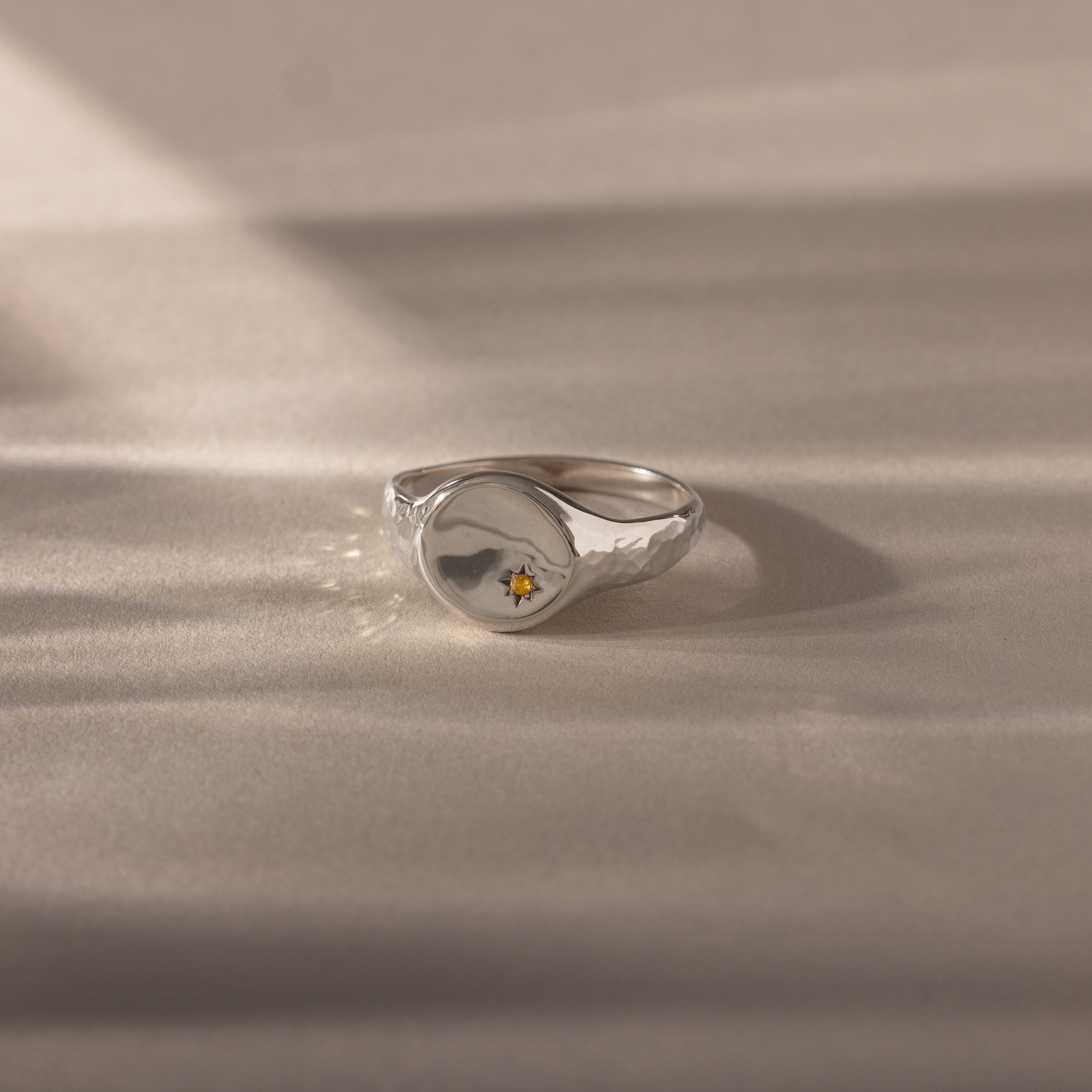 The North Star Topaz Signet Ring is a vintage-inspired silver ring featuring a small yellow gemstone at its center, beautifully displayed on a beige surface.