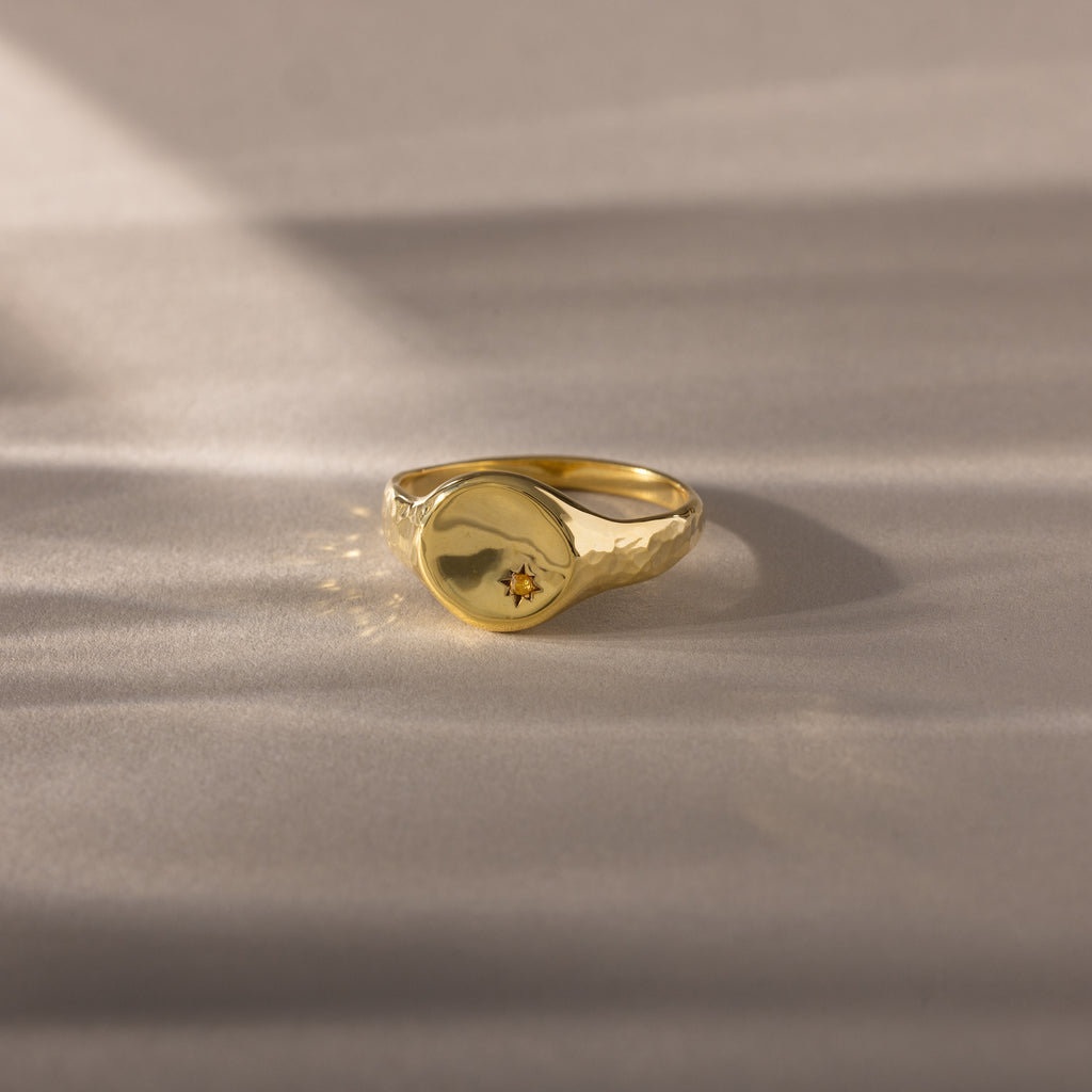 The North Star Birthstone Signet Ring in Topaz (November) features a small star-shaped engraving and is displayed on a soft, beige surface.