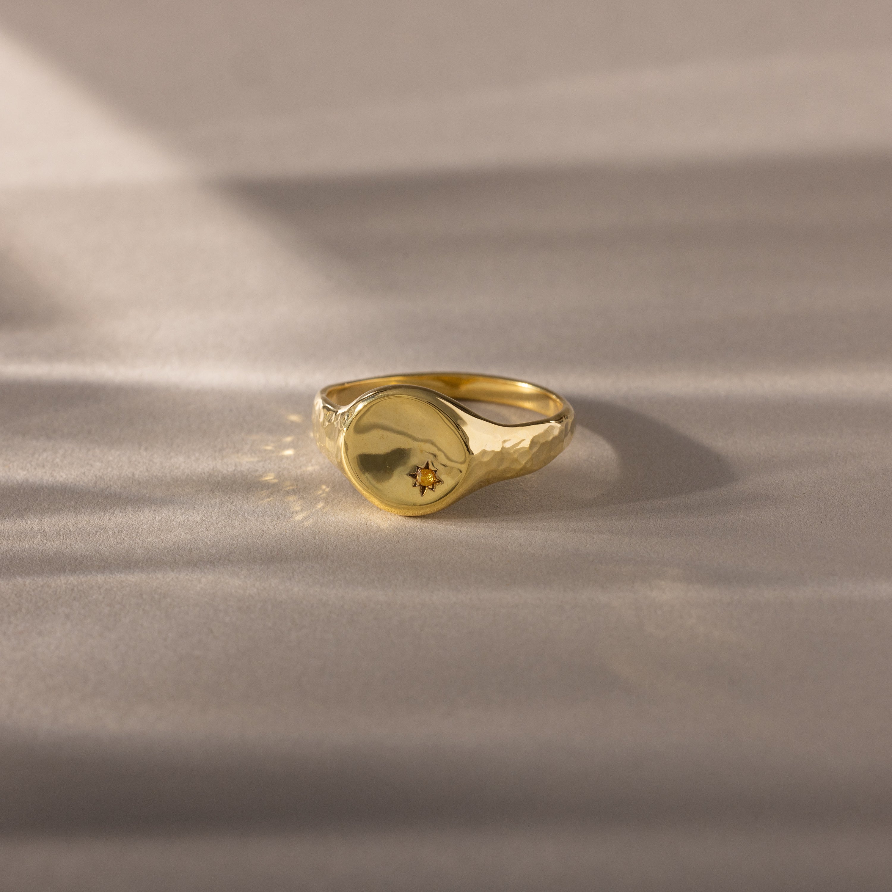 The North Star Birthstone Signet Ring in Topaz (November) features a small star-shaped engraving and is displayed on a soft, beige surface.