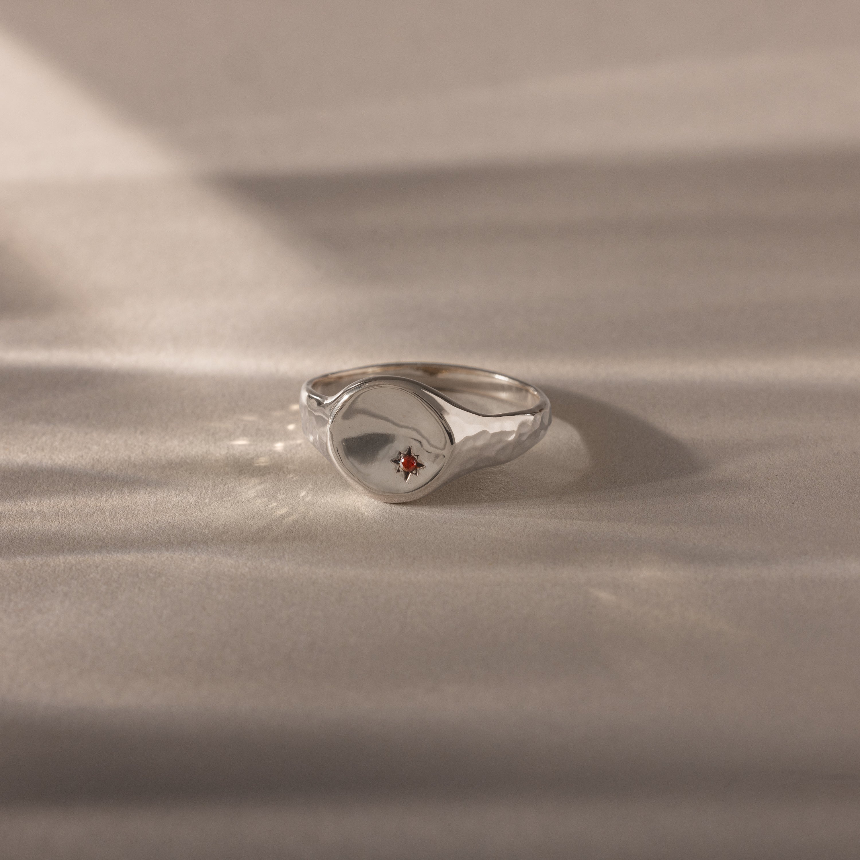 The North Star Garnet Signet Ring features an organic, wavy silver design with a small red garnet stone at the center, creating a vintage-inspired piece of symbolic jewelry displayed on a soft beige surface.