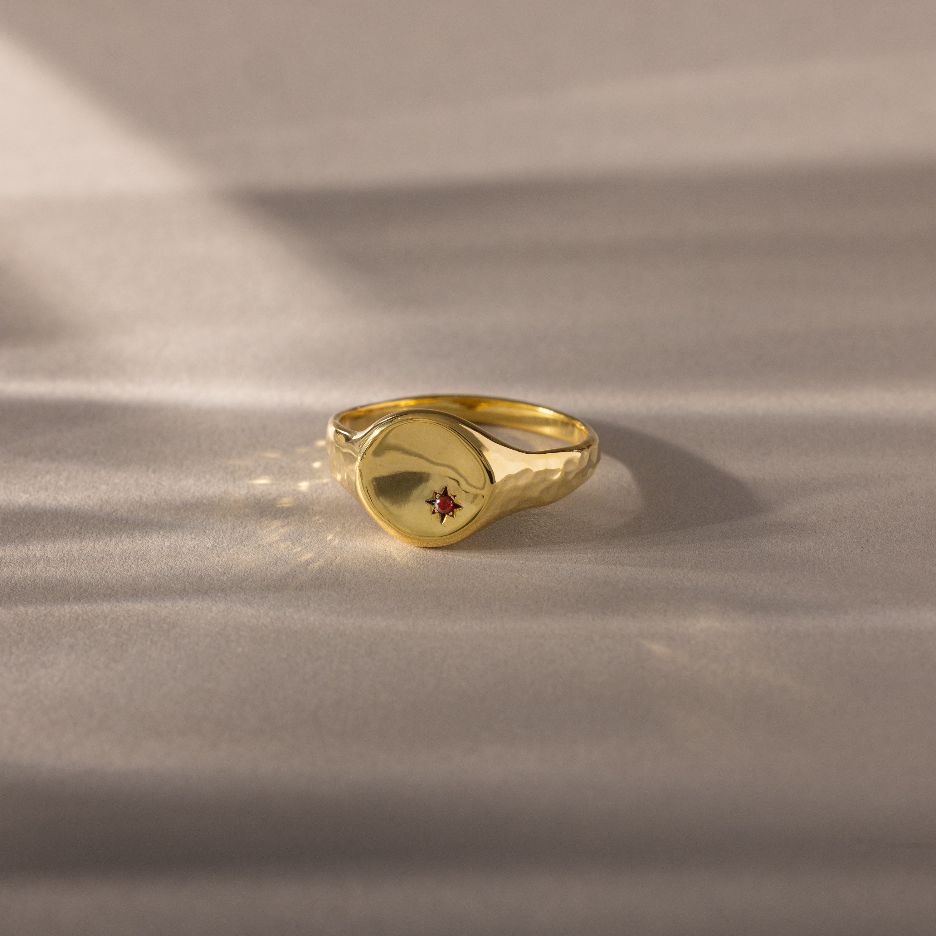 The North Star Birthstone Signet Ring in garnet, a symbolic gold piece for January birthdays, rests on a soft beige surface.